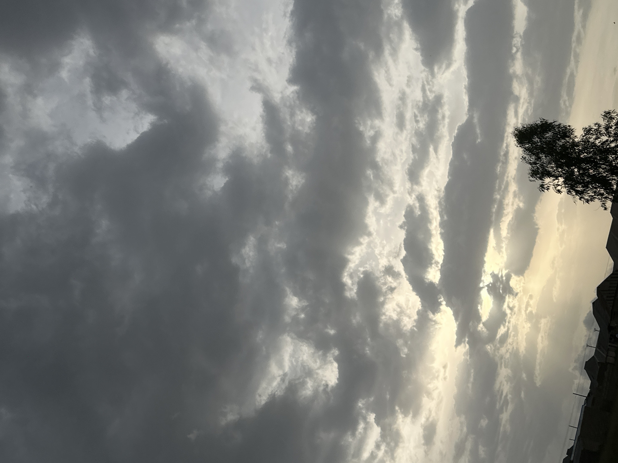 A low-angle, full shot captures the sky above a residential area in Durumi, Nigeria, during what appears to be sunset. The sky is dominated by thick, dark gray clouds with patches of lighter gray and white, suggesting an overcast or potentially rainy day. Sunlight filters through some of the thinner cloud layers, creating a diffused glow in the upper portion of the frame.
Along the bottom of the image, the rooftops of several buildings are visible, silhouetted against the brighter sky. These structures appear to be simple, residential houses with sloped roofs. A single, tall tree with dense foliage stands in the lower right quadrant of the image, its dark silhouette contrasting with the sky. The environment appears to be a relatively flat, urban or semi-urban landscape. No people are explicitly visible. The overall mood is somber and subdued due to the heavy cloud cover and muted lighting.