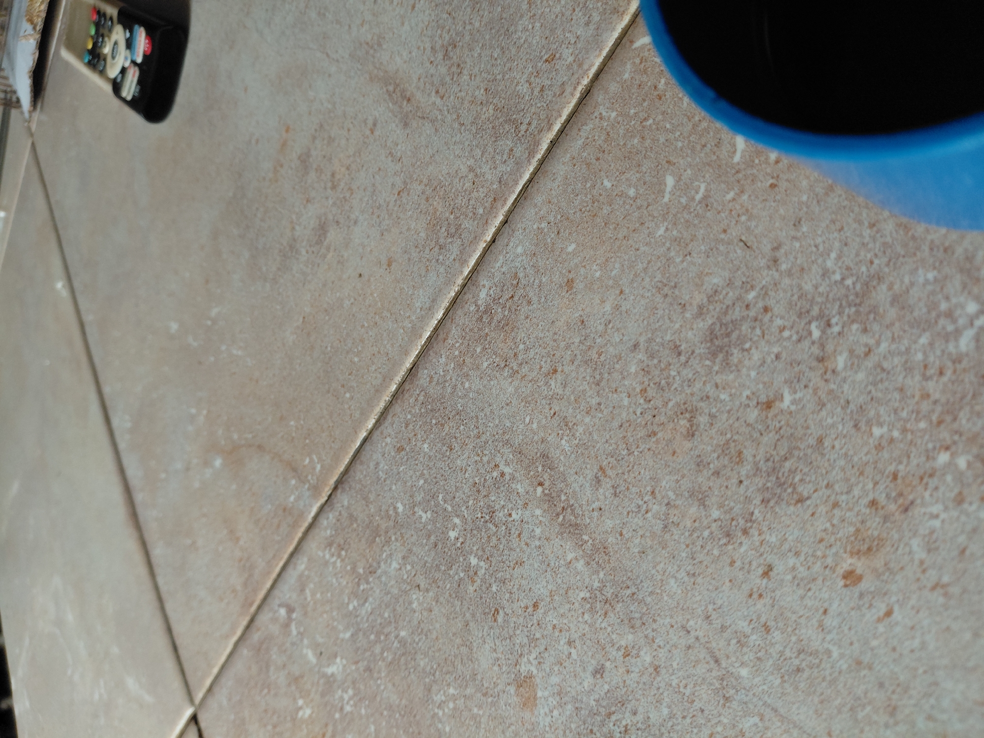 The image captures a close-up, high-angle view of a tiled surface, likely a floor or countertop, in what appears to be an indoor setting in Santa Ana, Venezuela. The tiles are rectangular, predominantly light beige or tan, with a textured, speckled pattern that mimics natural stone or ceramic. Visible grout lines separate the tiles, appearing slightly darker than the tile surface itself.
In the upper left corner, a black remote control is partially visible. It features several small, colorful buttons, including white, red, green, and blue, though no discernible text or brand name is legible. In the upper right corner, a portion of a blue cup or mug is visible, showing its bright blue exterior and a dark, possibly black, interior, suggesting it either contains a dark liquid or is empty.
No specific activity is taking place; the scene depicts a static arrangement of everyday objects on the tiled surface. The lighting is even, suggesting an indoor environment without strong directional shadows that would indicate a specific time of day. There are no people visible, and no emotions are conveyed.