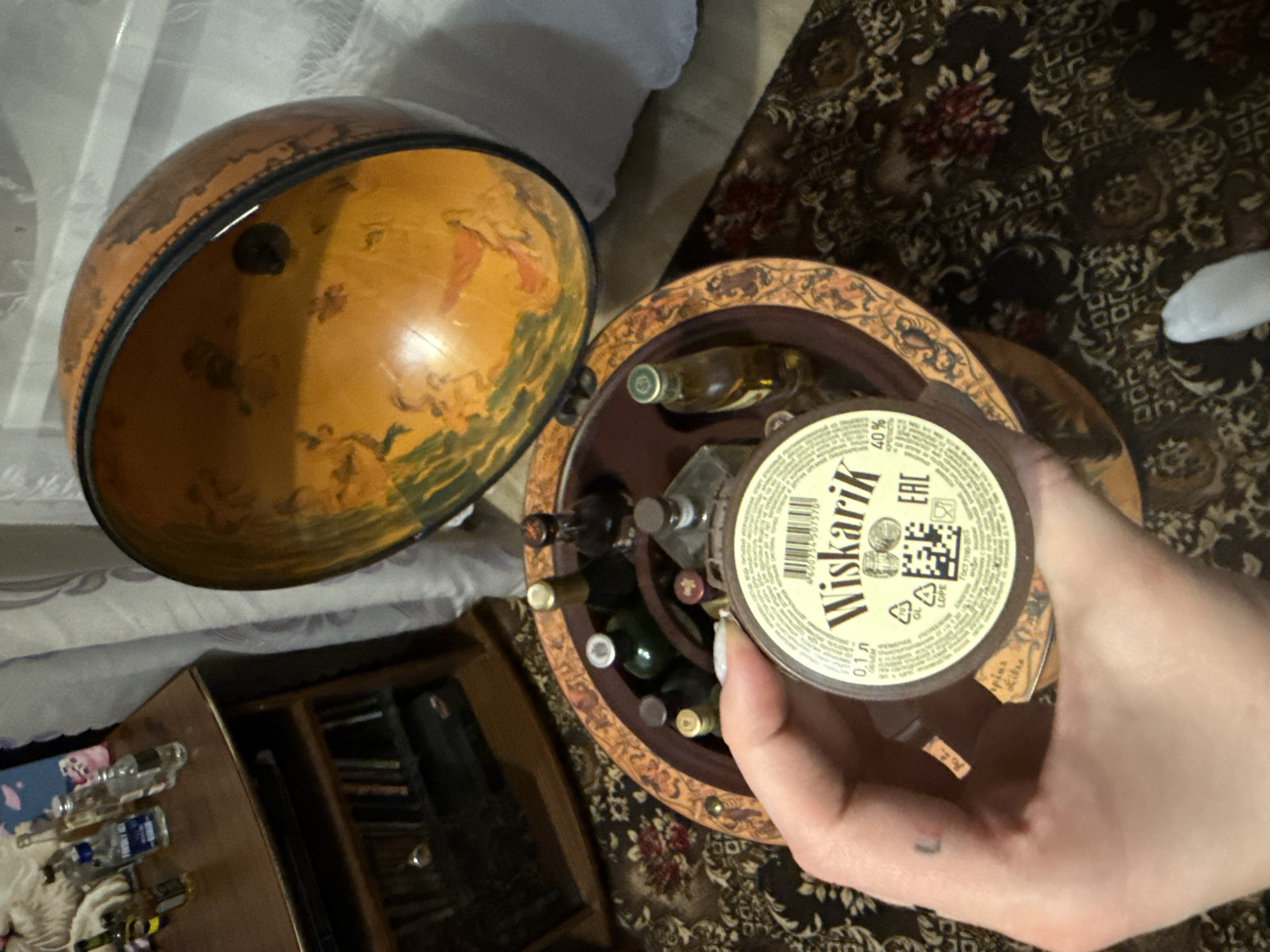 The image captures a top-down view of a person's right hand holding a small bottle, positioned above an open globe-shaped bar. The scene is set indoors, likely a home in Krapivka, Russia, with a dark, patterned rug covering the floor.
The main subjects include the hand and the bottle, an antique-style globe bar, and various miniature bottles. The globe bar is open, with its top half resting beside the base. The interior of the globe's top half features an aged map or artistic depiction with classical figures. The circular base of the globe bar serves as a rack, holding several small liquor bottles upright.
The bottle held by the hand has a white label with prominent black Cyrillic text that reads "Wiskarik." Below this, "40%" is clearly visible, indicating the alcohol content, and "0.1л" denotes a volume of 0.1 liters. Other smaller Cyrillic text, a QR code, a barcode (4606034507576), and an "EAC" (Eurasian Conformity) mark are also visible on the label. The hand displays light-colored nail polish and a small, faded tattoo on the side of a finger.
To the left of the globe bar, on a light-colored surface, several other small, clear glass bottles, some with blue labels, are arranged. Further left and partially obscured, a wooden cabinet or shelf unit containing books or other items is visible. The carpet underneath features intricate floral and geometric patterns in shades of red, white, and dark blue/black.
The lighting in the image is dim, suggesting it was taken during the evening or night under artificial light. No specific activity beyond holding the bottle and its proximity to the globe bar is evident.