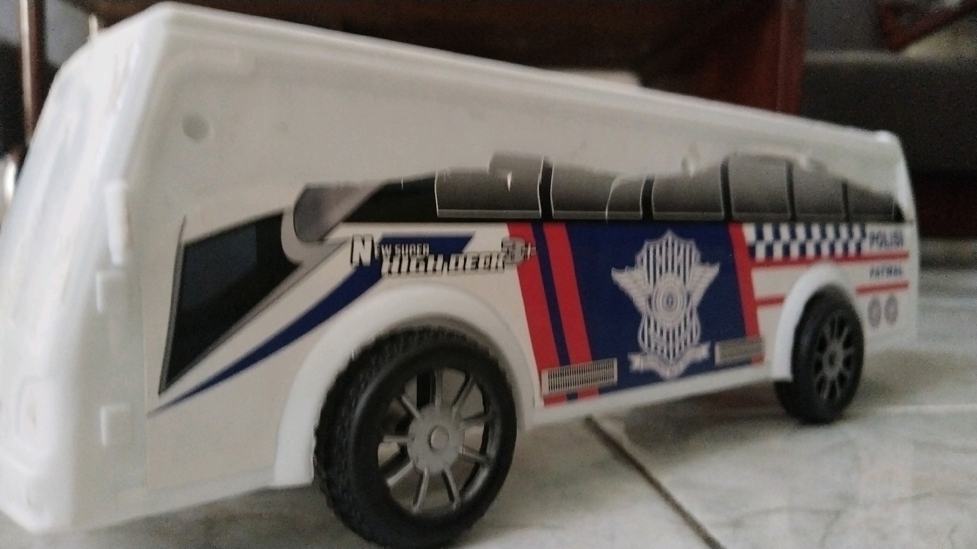A white toy bus, designed to resemble a police patrol vehicle, is positioned on a tiled surface. The bus features prominent markings, including a blue and red striped design, a police emblem, and text that appears to read "POLISI" and "PATWAL". The windows of the bus are represented by dark grey, segmented panels. The vehicle has black wheels with grey rims. The background is out of focus, but appears to be an indoor setting with wooden furniture. There is no visible indication of the specific location within Simo, Indonesia.