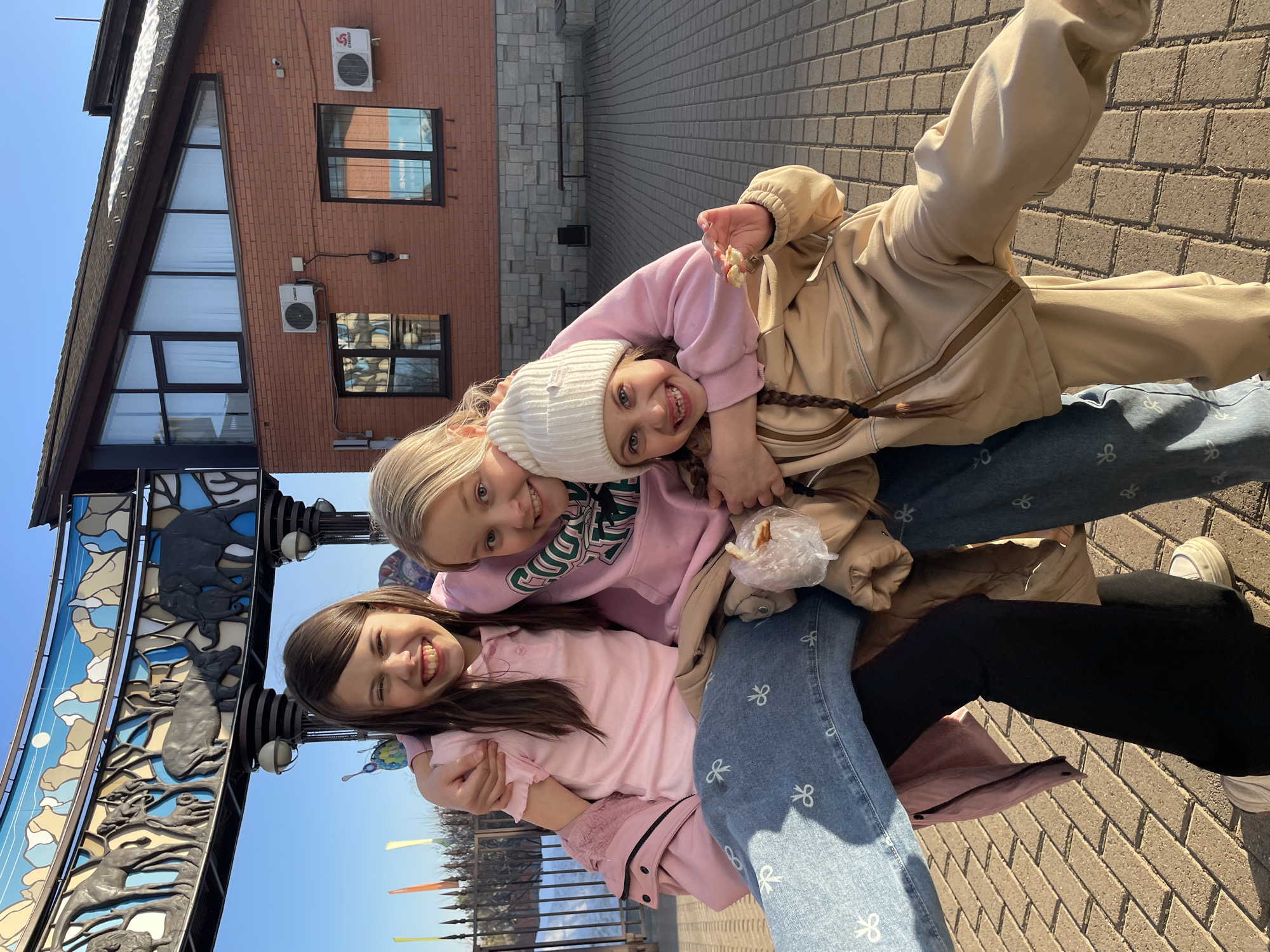Three smiling girls are piled on top of each other in front of a stylized metal gate depicting animals. The gate is located in Izhevsk, Russia, outside the Izhevsk Zoo. The girl on the bottom wears a pink shirt and light blue jeans with white bow patterns. The girl in the middle has on a pink sweatshirt with green lettering and a white beanie. The girl on top wears a beige jacket and jeans. All three girls are smiling at the camera. A brick building with multiple windows and two air conditioning units is visible in the background. The ground is paved with light brown bricks.