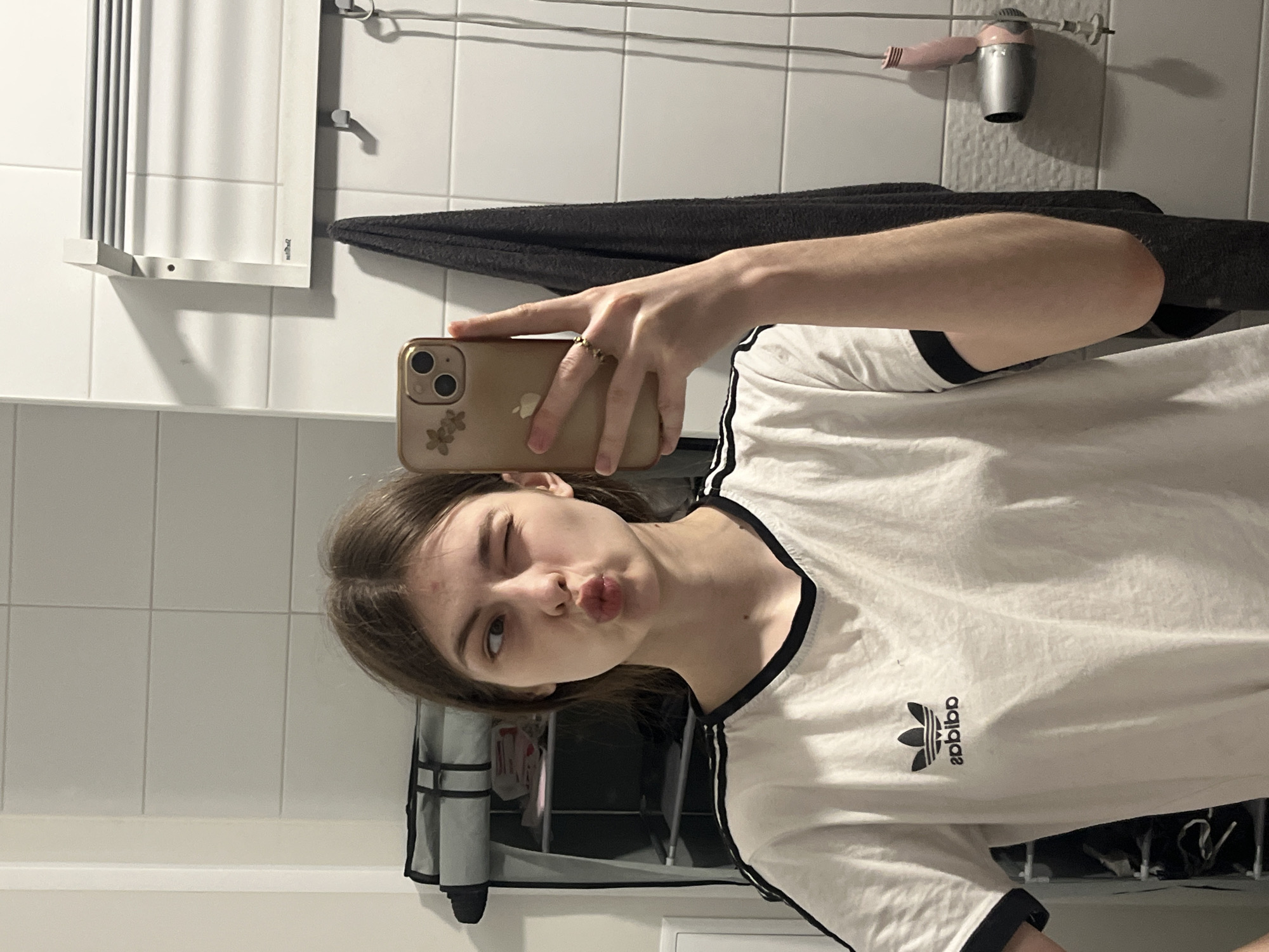 A young woman is taking a mirror selfie in what appears to be a bathroom. She has light brown hair, is winking with her right eye (from the viewer's perspective), and puckering her lips in a playful expression. She holds a gold-colored smartphone, possibly an iPhone, with a clear case adorned with floral stickers, in her right hand. A ring is visible on her right forefinger.
She is wearing a white t-shirt with black trim on the collar and short sleeves. The Adidas trefoil logo and the word "adidas" are visible on the front of her shirt, both appearing mirrored due to the reflection.
The background shows white tiled walls, characteristic of a bathroom. Visible fixtures include a white wall-mounted towel rack, a dark gray towel hanging below it, and a pink and silver/grey wall-mounted hairdryer positioned higher on the wall. Additional wall hooks are also present. A storage unit with compartments can be partially seen reflected behind her, further indicating an indoor, functional space. The lighting is artificial, suggesting an indoor setting without external light cues.