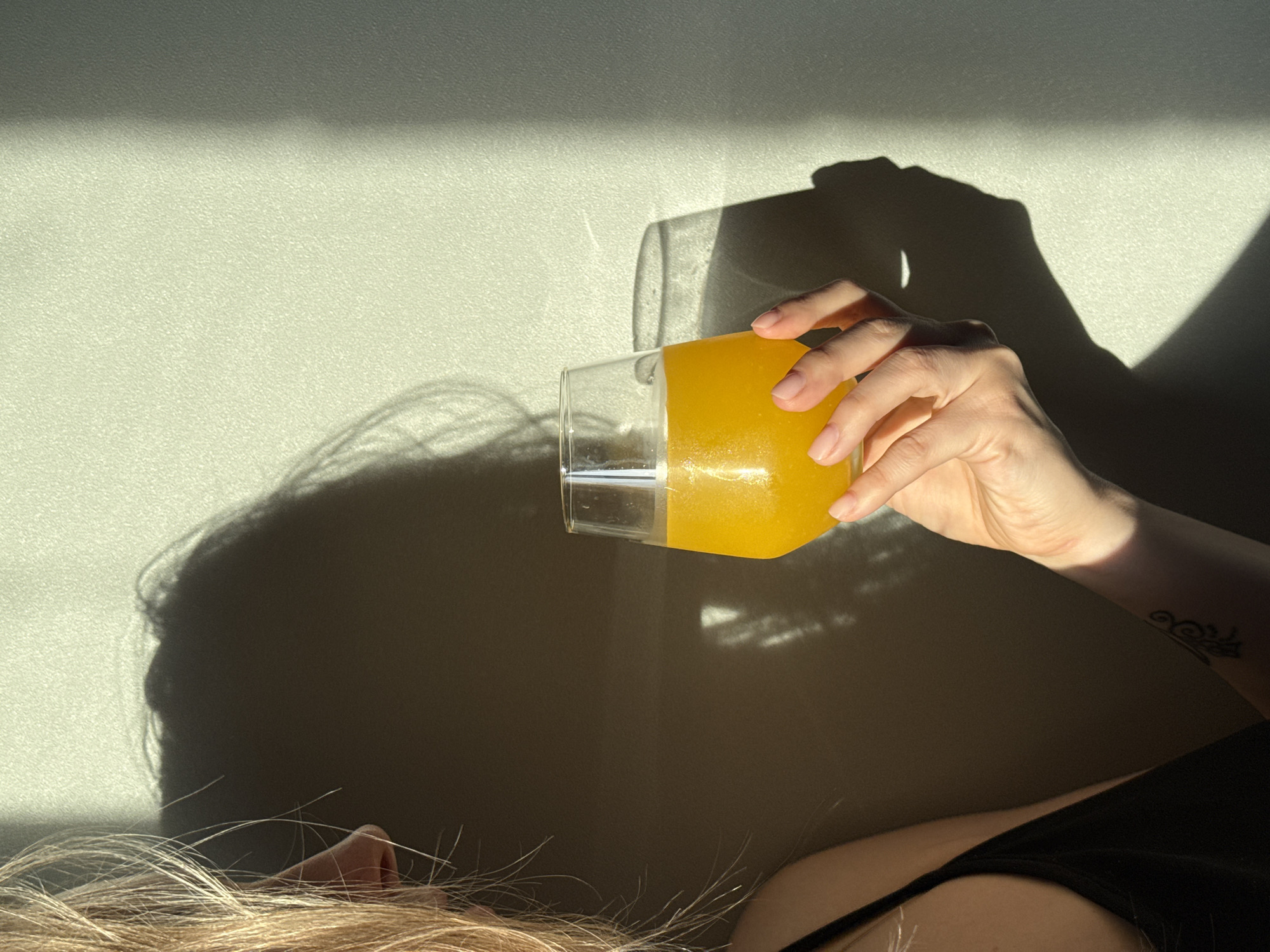 A woman with light blonde hair is lying down, holding a glass of orange juice. Her right hand is visible, with a tattoo on her arm. She is wearing a black tank top. The glass casts a shadow on the wall behind her, along with the shadow of her head. The wall is light-colored and appears to be textured. The light is strong and creates defined shadows. The location is unknown, not verifiable as Chaikovsky, Russia.
