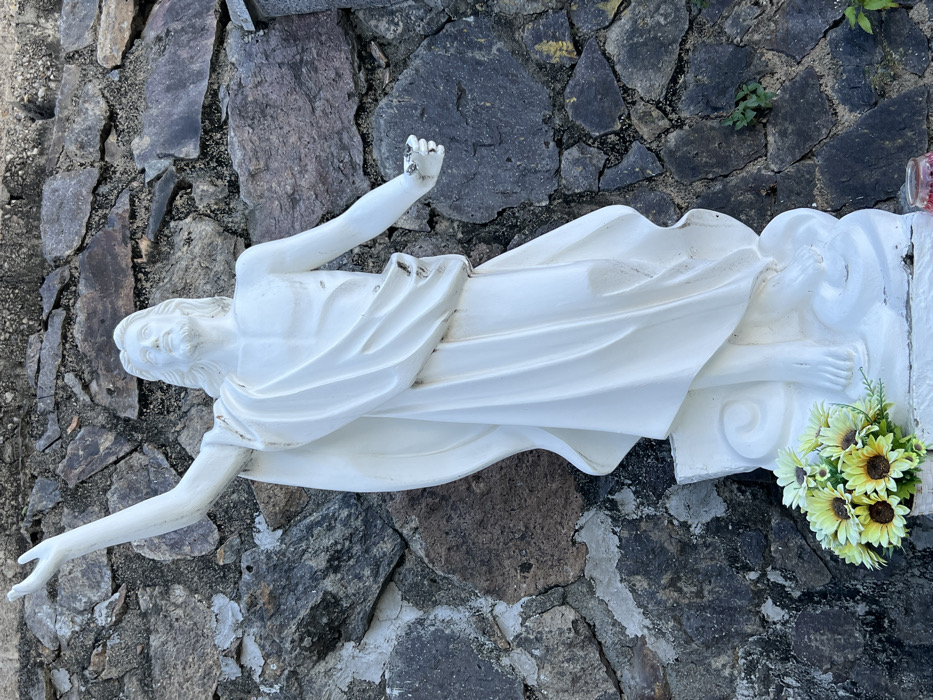A full-length white statue of Jesus Christ stands against a rough-hewn stone wall, likely located at or near Nha Trang Cathedral (Christ the King Cathedral) in Nha Trang, Vietnam. The statue depicts Jesus with a beard and long hair, clad in a flowing robe draped over his left shoulder and lower body. His left arm is raised vertically with an open hand, while his right arm extends forward and slightly outward, also with an open hand. His head is tilted upward, and his gaze is directed skyward. The statue rests on a white, cloud-like sculptural base, displaying minor discoloration and weathering. At the base of the statue, on the left, a small bouquet of artificial sunflowers and white daisies with green foliage is present. A red object, possibly a votive candle in a glass holder, is partially visible in the lower right foreground. The background consists of an irregular wall constructed from various sizes of dark grey and brown natural stones, with small patches of green vegetation growing from its crevices. The scene is brightly illuminated, indicating daylight conditions.