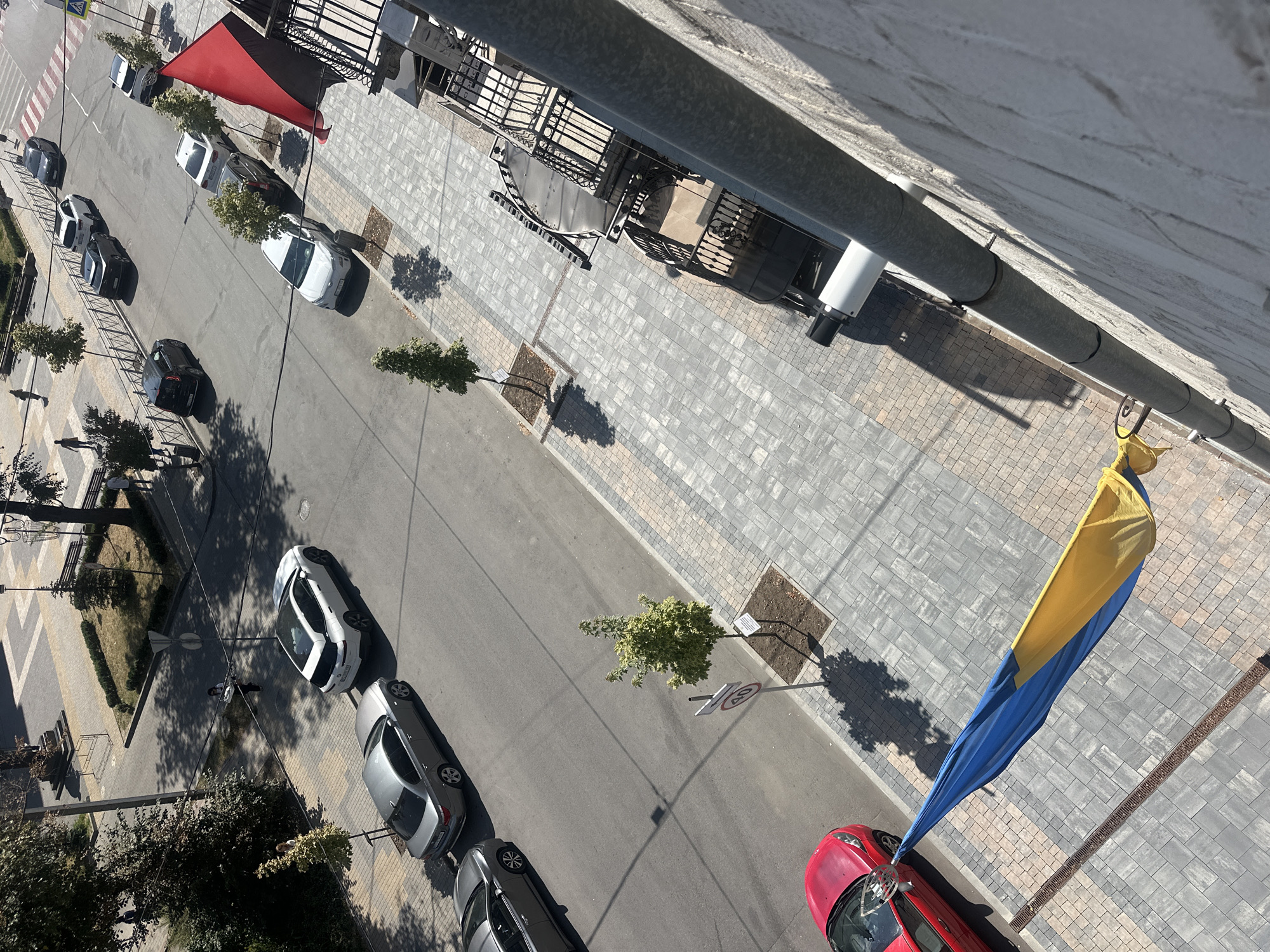 An overhead shot captures a street in Ternopil, Ukraine, bathed in sunlight. A variety of cars, including silver sedans, a white SUV, a black hatchback, and a red sports car, are parked or driving on the asphalt road. Adjacent to the road is a paved pedestrian walkway with trees lining it, casting dappled shadows. Two flags are visible: a red and black flag flies from a balcony on the right side of the image, and a blue and yellow Ukrainian flag drapes across the walkway in the foreground, partially obscuring a red car. Several people are present, walking on the sidewalk and the street. Buildings with balconies and architectural details are visible on the right side of the frame. The environment suggests an urban setting on a clear day.