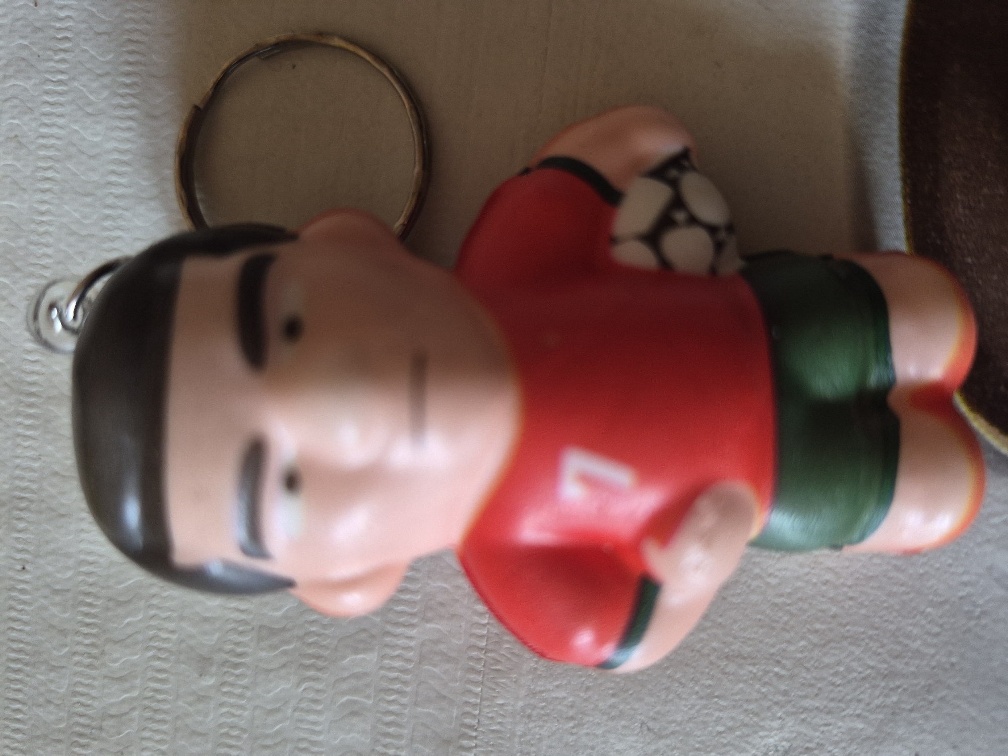 The image presents a close-up of a plastic figurine keychain, oriented horizontally, lying on a textured, light-colored surface. The figurine portrays a stylized male figure, likely a footballer, with a light-skinned head featuring black hair, distinct black eyebrows, and small dot-like eyes. He is dressed in a bright red jersey, which has a blurry white number, appearing to be '7', on its front. He also wears green shorts and holds a small white football with black patterns under his right arm. A silver metal keyring is attached to the top of the figurine's head. In the background, a larger, gold-colored metal ring is partially visible, along with a darker, possibly brown, surface on the right side. The foreground surface beneath the figurine exhibits a repeating wavy or ribbed pattern. The overall image is slightly out of focus, particularly the figurine's face and the number on its jersey. No activity is depicted; it is a static shot of the object. While the image itself provides no geographical markers, its context places it in Yerevan, Armenia.