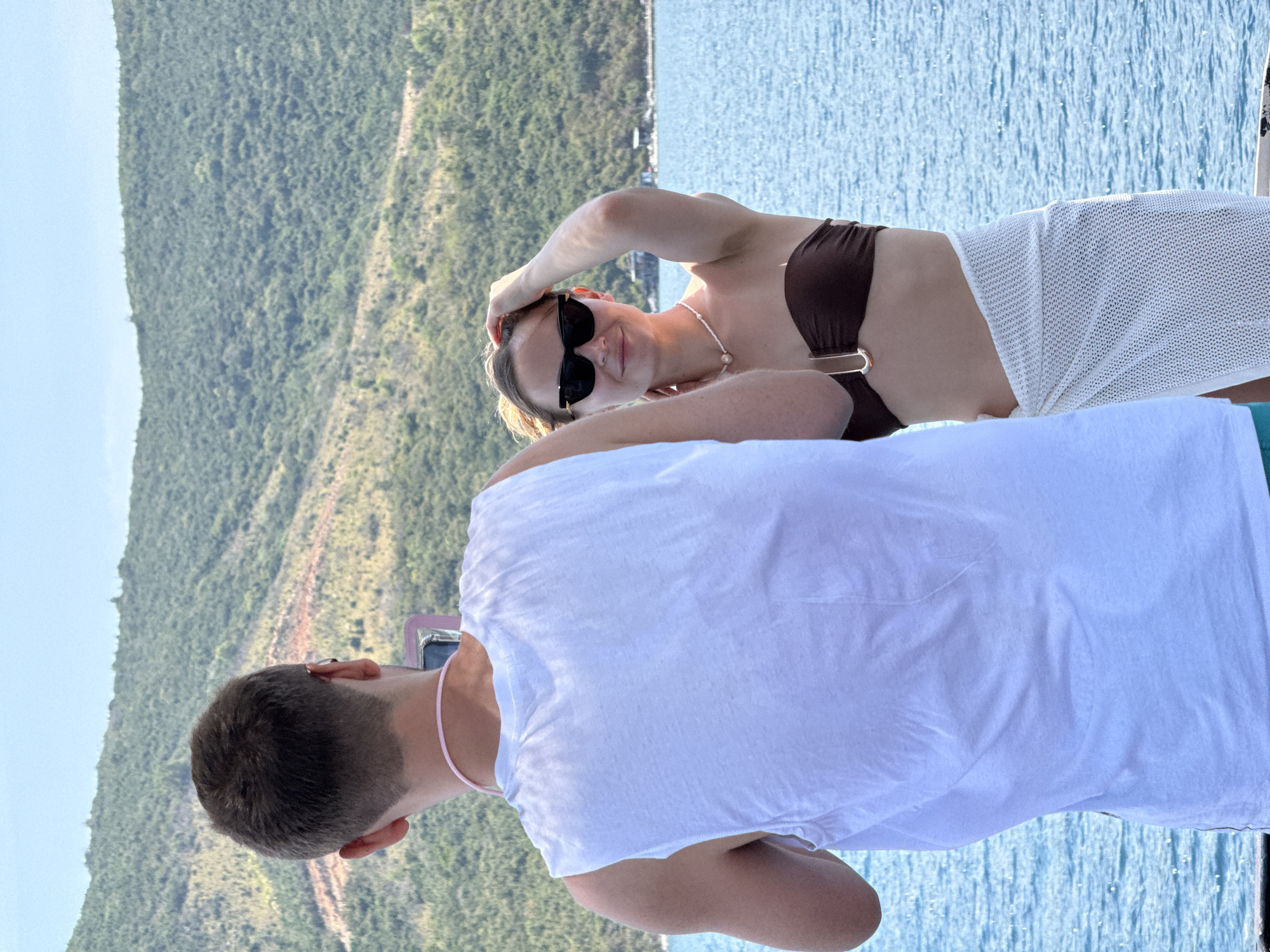 A rotated shot shows a man and woman outdoors, likely on a boat, with a coastline in the background. The woman is lying down, wearing a brown bikini top, a white mesh cover-up, and sunglasses. She has one hand on her head. The man, facing away from the camera, wears a white shirt and a pink necklace. Water is visible in the background, and the coastline features greenery and possibly some structures. The location is in Nha Trang Ward, Vietnam.