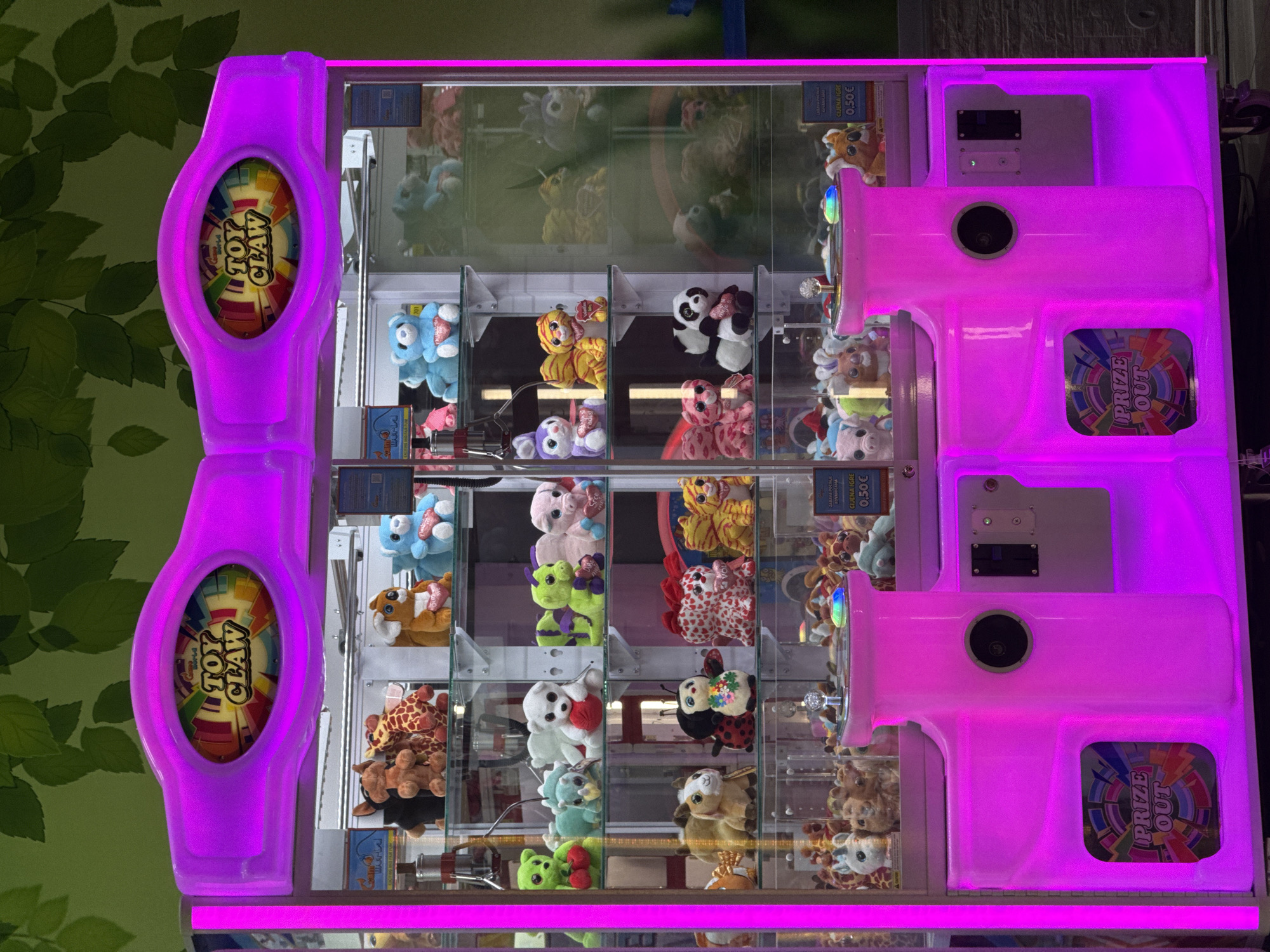 This image captures a vibrant, brightly lit claw machine, likely situated in an indoor entertainment venue such as an arcade or family center. The machine is adorned with striking neon purple lighting along its edges and control panels, creating a lively atmosphere.
The primary subjects are the numerous plush toys displayed within the clear glass compartments of the machine. These include a variety of stuffed animals such as blue bears, pandas, tigers, pigs, green dragons, ladybugs, and giraffes, neatly arranged in rows, waiting to be won. The machine appears to be a multi-level or double-sided unit, with two distinct play areas visible.
No people are present or actively playing the game, but the machine is fully stocked and ready for use. The glowing purple controls feature a central, colorful emblem that reads "TOY CLAW."
Notable details include several informational signs. On the top left, text states "ZABAVA POČINJE KOVANICAMA" (Fun starts with coins) and "CIJENA IGRE 0,50€" (Price of game 0.50€), indicating the cost per play. Similar price signs are visible elsewhere on the machine. Below the control panels, a slot marked "PRIZE OUT" with a colorful, abstract design indicates where prizes are dispensed. The background behind the machine features a green wall decorated with leafy patterns. The image is oriented vertically, with the machine appearing on its side.