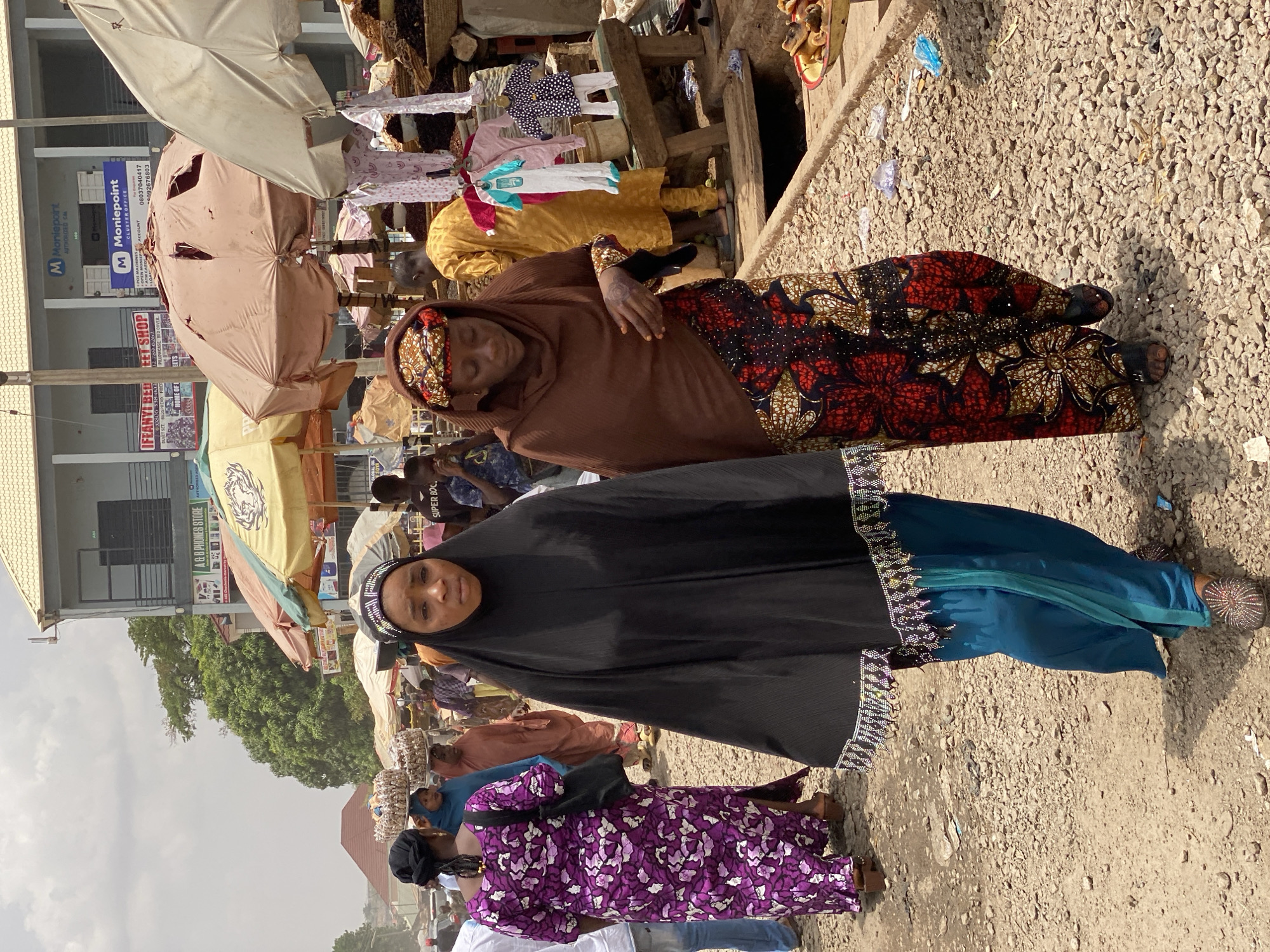 The image shows a bustling street scene in Maiburiji, Nigeria. Two women are prominently featured. The first woman wears a black head covering with a decorative border, a blue undergarment, and ornamented shoes. Behind her, another woman wears a patterned brown head covering and dress of similar pattern. Both stand on a dirt and gravel street. In the background, a store sign reading "A & B Phones Store", "Moniepoint", and "Ifeanyi Bed Sheet Shop" can be seen, alongside umbrellas. Clothes hang above the stalls and buildings. Other figures are present, but are further in the background.