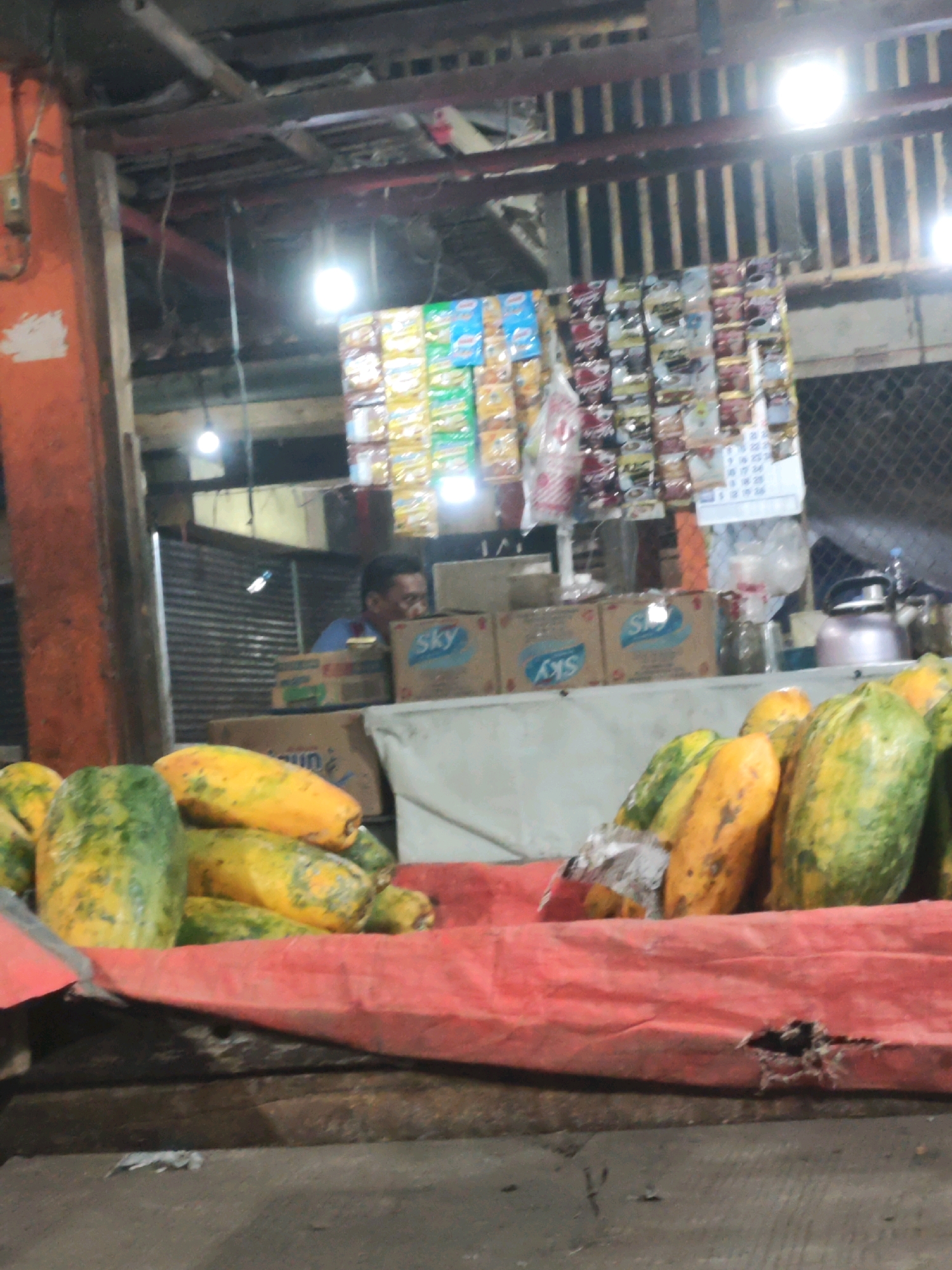 This is a medium-shot, eye-level photograph of a fruit stand in Karawang, Indonesia. The stand is located outdoors, likely in a market, with a corrugated metal roof supported by wooden beams overhead. String lights illuminate the scene, casting a warm glow on the produce. In the foreground, a pile of ripe papayas is displayed on a bright red tarp. The papayas vary in color from green to yellow and orange, indicating different stages of ripeness. Behind the fruit, several cardboard boxes are stacked, with some bearing the "Sky" logo. A man, presumed to be the vendor, is partially visible in the background, looking down at the fruit. Shelves above the boxes are stocked with various packaged goods, possibly snacks or instant food items, with a small calendar attached to the wall. The overall atmosphere suggests a bustling, everyday scene at a local market.