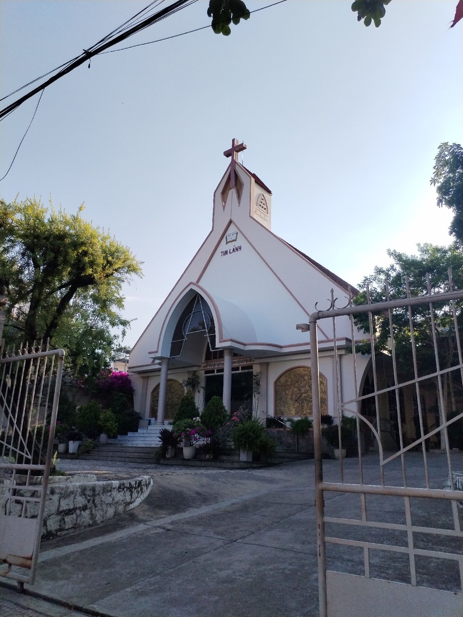A church stands in Nha Trang Ward, Vietnam. The church is light-colored with a cross on top. A sign is visible on the front of the church. A gate surrounds the yard in front of the church. Trees and potted plants are planted around the building. The sky is blue and clear.