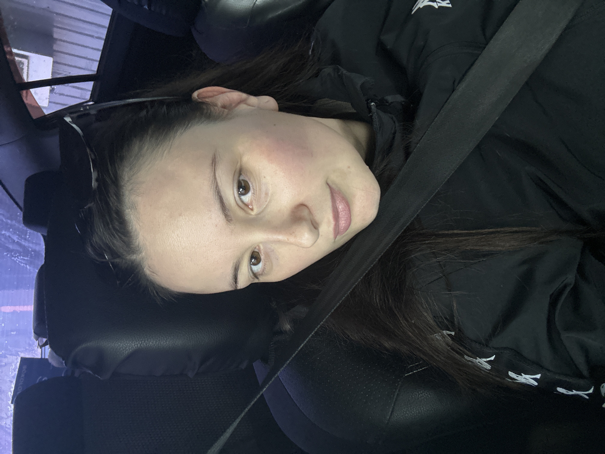 A woman with dark hair is seated in what appears to be the passenger seat of a car. She is wearing a black jacket and a seatbelt is fastened across her chest. Her head is resting against the headrest. A pair of glasses are propped up in her hair. The location, Chaikovsky, Russia, is not evident from the visible contents of the image.