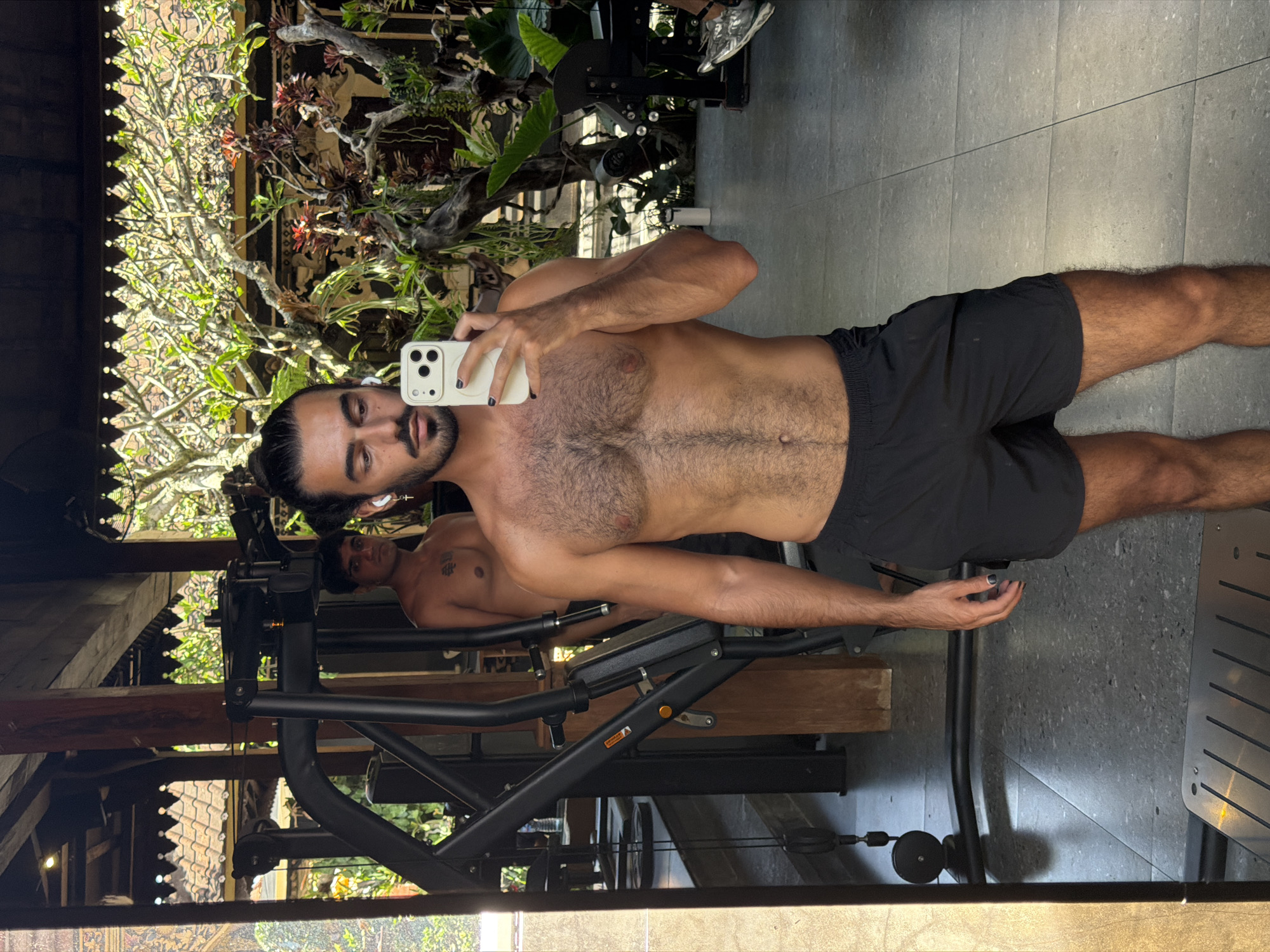 This media file features a shirtless man taking a mirror selfie in what appears to be a gym or fitness area. He has dark hair pulled back and is wearing black athletic shorts. His chest and abdomen are visible, and he has body hair. He is holding a white smartphone in his right hand, and his left hand is resting on his thigh.
In the background, through the reflection in the mirror, we can see gym equipment, including a weight machine with black metal framing and wooden accents. There are also lush green plants and trees visible, suggesting an outdoor or semi-outdoor setting, possibly in a tropical location. The overall impression is of a relaxed, possibly humid environment, given the greenery.
Another person is visible in the background reflection, seated on a weight machine and looking towards the camera. This second individual also appears to be shirtless.
The lighting suggests it is daytime, with natural light filtering in. The man's expression is neutral, and he appears to be focused on taking the selfie. There is no visible text within the image. The overall scene evokes a sense of fitness and well-being in a natural, possibly exotic setting.