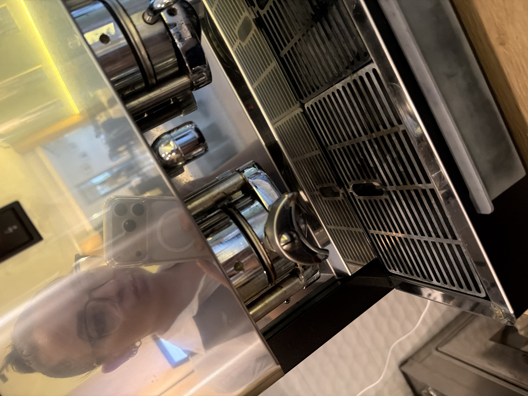 A woman, visible in a reflection, is taking a selfie with her phone in front of a stainless steel espresso machine. She is wearing glasses and has her hair pulled back in a bun. The espresso machine has a drip tray with a metal grate and various buttons and spouts. The scene appears to be indoors, possibly in a kitchen or cafe, with a wooden countertop visible in the foreground. There is no indication of the location being in Otranto, Italy.