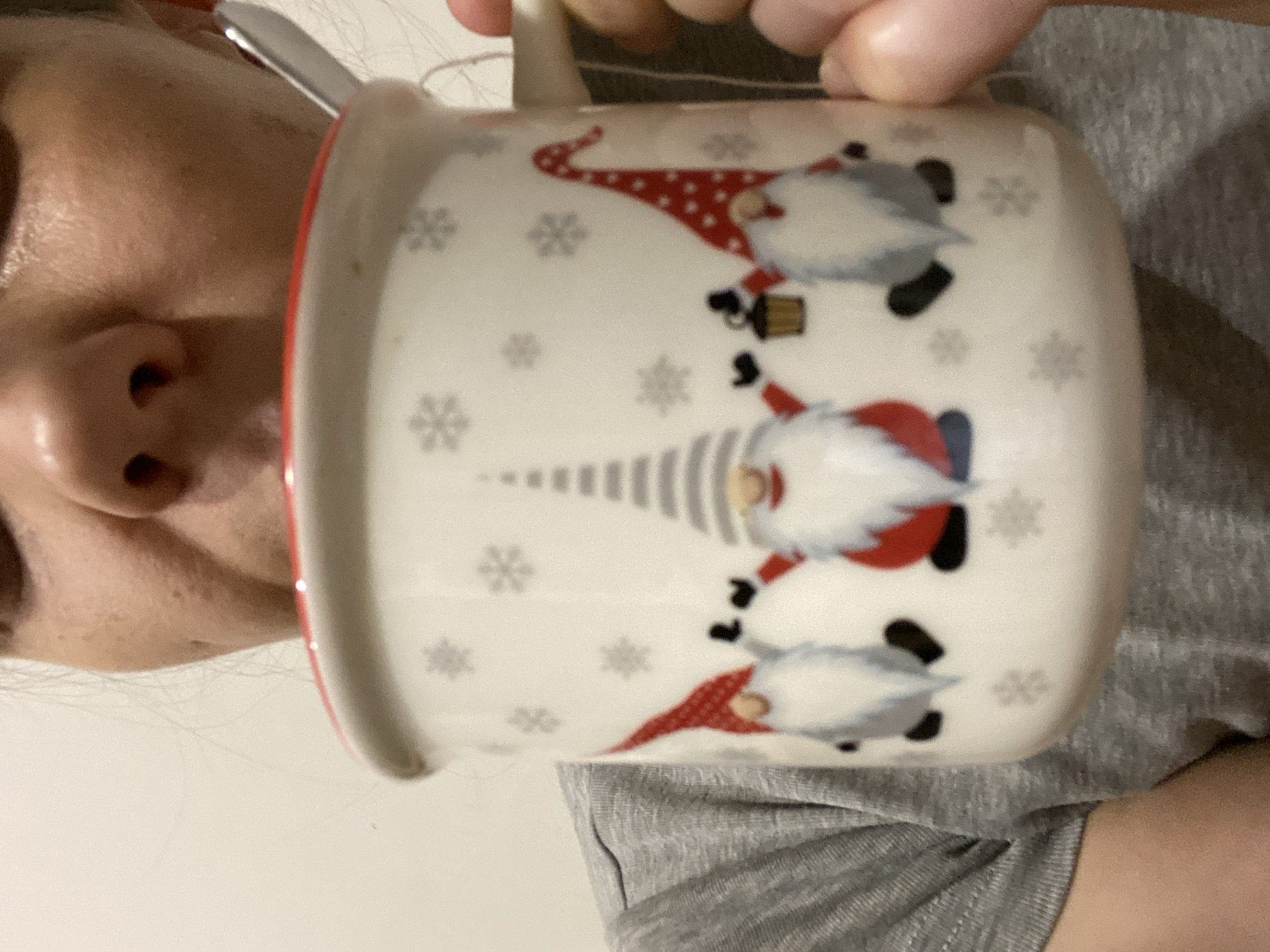 The image depicts an individual partially obscured by a mug, observed in an indoor setting within the city of Chaikovsky, Russia. The person, from the nose down to the upper chest, holds a white ceramic mug with a red rim to their mouth, actively engaged in drinking. A metallic spoon is visible inside the mug. The person is wearing a grey, short-sleeved t-shirt. A white earbud wire is visible near the individual's right ear. The mug features a repeating pattern of grey snowflakes and three distinct gnomes: one on the left with a red hat, a central gnome with a grey and white striped hat, and a gnome on the right with a red polka-dotted hat holding a dark lantern. The background is a plain, light-colored wall.
