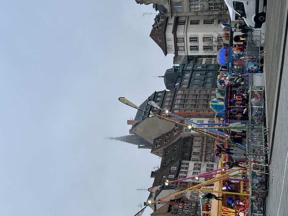 The image displays an outdoor fairground set up in a public square in Strasbourg, France, likely near Place du Château, with the spire of Cathédrale Notre-Dame de Strasbourg partially visible through an overcast, hazy sky in the background.
In the foreground and midground, several amusement attractions are present on a dark paved surface, enclosed by metal crowd control barriers adorned with comic book hero artwork. To the left, a bungee trampoline ride features multi-colored frames (yellow, pink, blue) with integrated lighting. Two individuals are visible in harnesses on this ride; one is suspended in mid-air, and another is positioned lower down. Two tall, narrow yellow banners are attached to the trampoline structure. To the right of the bungee trampoline, a carousel-style ride, also illuminated with blue lights, is visible with several people seated on it. Its base panels also display comic book hero themes. Red and white striped tape lies on the ground near the barriers. On the far right, a white van is parked.
The background consists of a dense arrangement of multi-story European-style buildings, exhibiting varied architectural details, rooflines, and facade colors, ranging from white and beige to brick-red and dark teal. Above these buildings, the intricate, dark Gothic spire of Strasbourg Cathedral rises into the dull grey sky, its upper section somewhat obscured by atmospheric haze or fog.