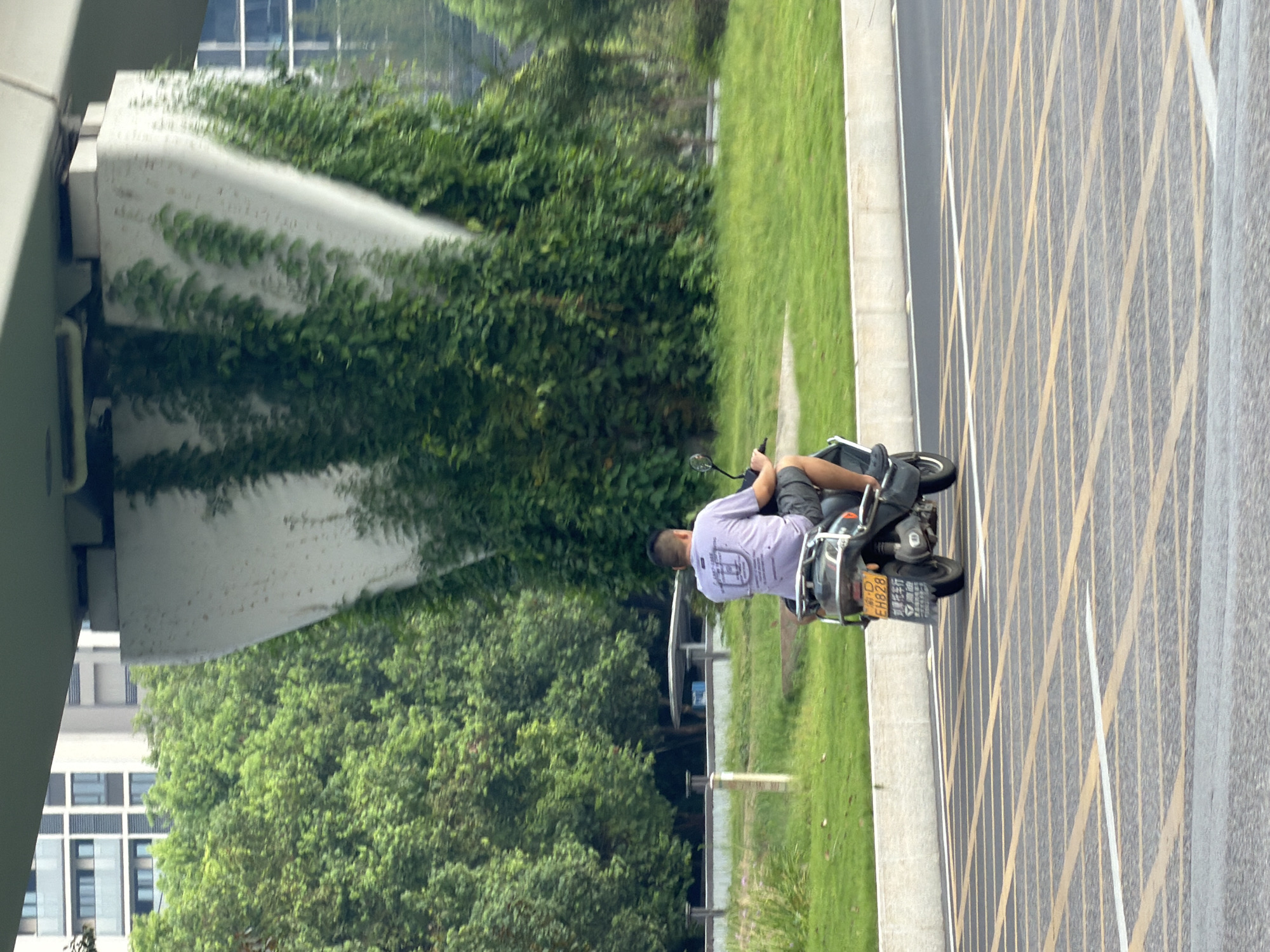 A man rides a motorcycle on a road in Jiangjin, China. The man is wearing a light purple t-shirt and dark shorts. The motorcycle is black and has a license plate that reads "EH828". The road has yellow and white stripes painted on it. In the background, there are trees, buildings, and a large concrete structure covered in vines. The overall scene appears to be an urban or suburban environment.
