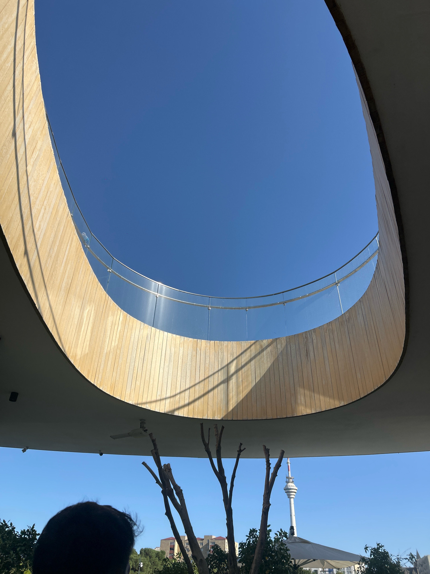 An outdoor, eye-level, wide shot depicts a view looking upwards towards a circular opening in a modern building structure. The opening is framed by a wooden facade on the left and a gray concrete overhang on the right. A clear blue sky fills the upper portion of the frame, with a thin, reflective glass barrier with metal railings encircling the opening. Sunlight casts striped shadows from the railing onto the wooden facade. Below the overhang, the bare branches of a tree are visible, along with lush green foliage. In the distance, a tall, slender broadcast tower, characteristic of Sabail Raion, Azerbaijan, stands against the blue sky. A portion of a person's dark hair is visible in the lower left corner, and a white umbrella is partially visible in the lower right. The scene suggests a contemporary architectural design set within an urban landscape.