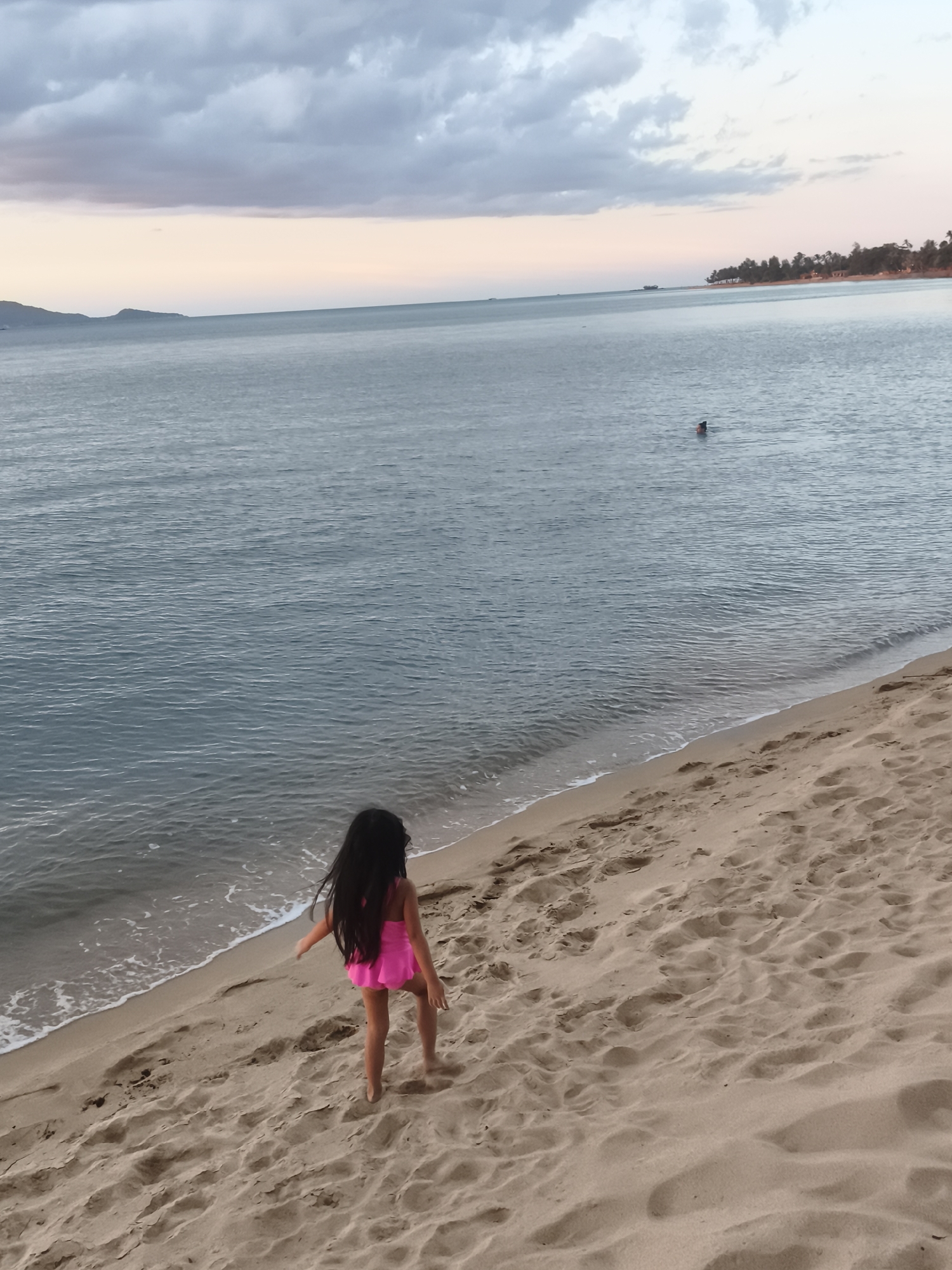 A young girl with long dark hair, wearing a bright pink swimsuit, walks along a sandy beach towards the gentle waves of the ocean. The sand is a light tan color and shows footprints from previous visitors. The ocean is a calm, deep blue, with small waves lapping at the shore. In the distance, a person is swimming in the water. The sky above is a mix of soft gray clouds and a pale pink and orange hue, suggesting it is either sunrise or sunset. The background shows a distant coastline with trees and a faint silhouette of hills, possibly indicating a tropical location like Baan Bo Phut, Thailand. The overall mood is peaceful and serene, capturing a quiet moment at the beach. No visible text is present in the image.