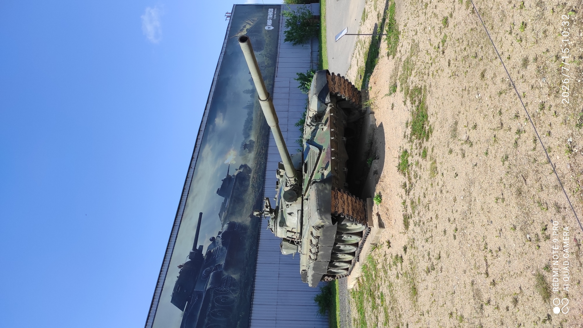 A green tank is parked outdoors on a sunny day in front of a building with a large mural depicting tanks. The tank is on a raised dirt platform, and it is facing to the right of the image. The mural behind the tank shows several tanks, one of which is firing its cannon. The building has a grey, corrugated metal exterior. The sky is a clear blue with a few small clouds. The ground in front of the tank is covered in sand and sparse vegetation. A small sign is visible to the right of the tank. The text on the mural in the upper right corner appears to be in Russian and reads "МИР ТАНКОВ". The timestamp in the bottom right corner reads "2026/7/15 10:32". The text in the bottom left corner reads "REDMI NOTE 9 PRO AI QUAD CAMERA". This appears to be an exhibit at a tank museum.