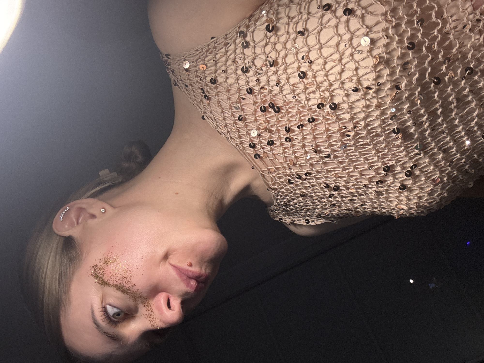 The image, rotated 90 degrees counter-clockwise, depicts a young adult woman in an indoor setting, possibly in Chaikovsky, Russia. Her face, in the lower left of the frame, is looking upwards and slightly to the left, with light-colored eyes. Gold-colored glitter is applied across her left cheek and nose, suggesting a festive or artistic makeup choice. She has multiple ear piercings on her left ear, including a cluster of small gems and a single stud. Her hair, which appears light in color, is pulled back.
She is wearing a light-colored, possibly beige or nude, top with a distinct open-weave, crocheted, or net-like texture, adorned with numerous small, reflective sequins. The top covers her shoulder and upper chest. A bright light source is visible in the upper left of the frame, illuminating the woman's face and the sequined top. The background is largely dark and indistinct, with a few faint light reflections visible in the distance on the right, but no specific objects or text can be identified. The low lighting and glitter suggest an evening event or a styled photoshoot.