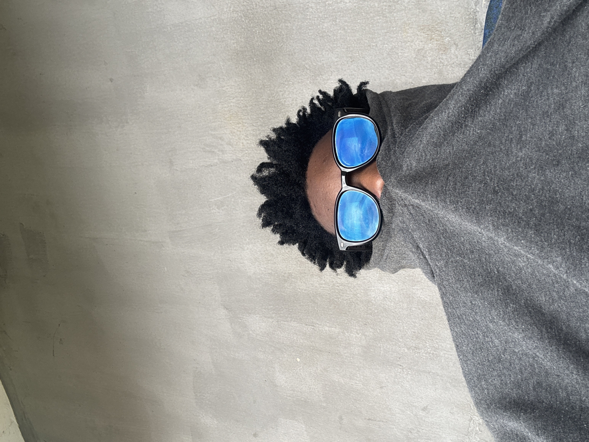A person is photographed from the chest up, against a plain, textured gray wall that appears to be a building exterior. The individual is wearing dark-rimmed sunglasses with bright blue lenses that reflect the sky. A dark gray hooded garment is pulled up to cover the lower half of the person's face, obscuring their nose and mouth, and a portion of their curly dark hair is visible above the sunglasses. The overall scene is minimalist, with the focus on the individual and their striking eyewear. The specific location within Ihiagwa, Nigeria, cannot be determined from the visual information provided.