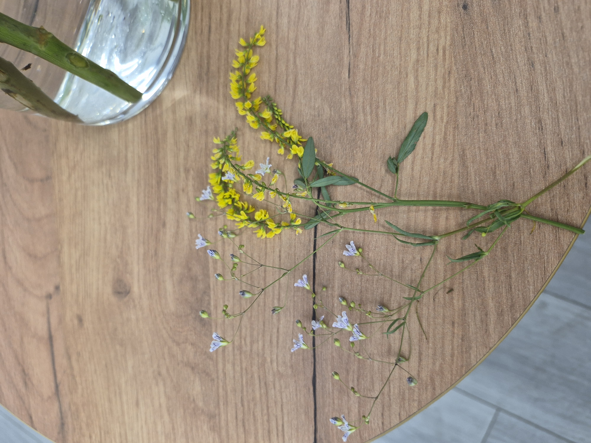 The image depicts a close-up, overhead view of a wooden tabletop. Two delicate bouquets of wildflowers are arranged on the surface. One bouquet features clusters of small yellow flowers, while the other has slender stems adorned with tiny light purple and white blossoms. In the upper right corner, a portion of a clear glass vase filled with water is visible, containing two green plant stems. The background consists of the textured wooden surface of the table, with a subtle hint of a blue-grey tiled floor visible at the bottom left edge of the frame. The scene is static and does not show any people or animals. The lighting suggests an indoor setting, possibly a home or a cafe.