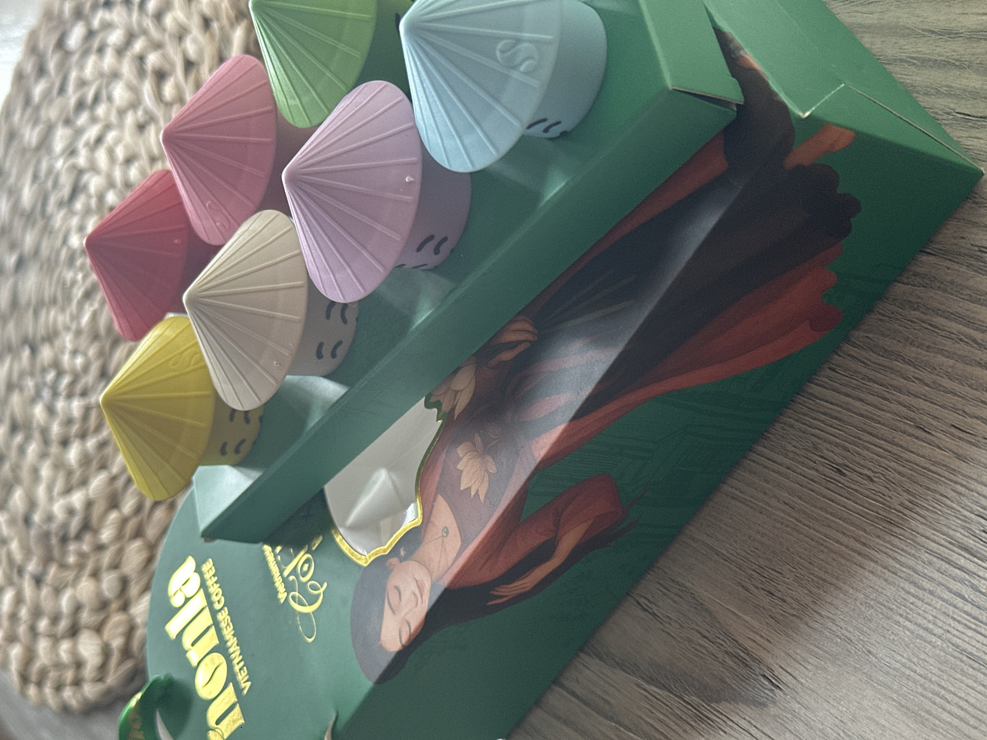A tilted, medium-shot depicts a green box, partially open, displaying an illustration of a woman in a flowing reddish robe. On top of the box, a collection of small, colorful, conical objects resembling hats or toys are arranged. The colors include green, light blue, lavender, beige, red, and yellow. The text "Nomla VIETNAMESE COFFEE" is visible on the box. The background includes a woven mat and a wooden surface. The location is not discernible from the image.