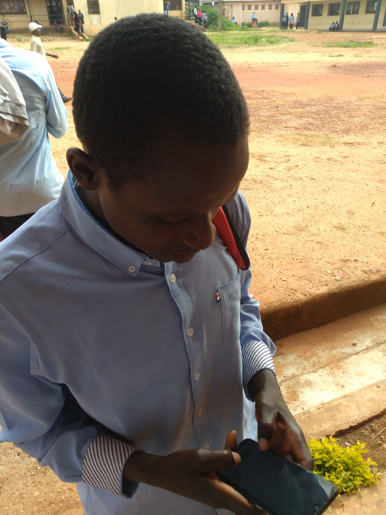 An individual, likely a young male, is the primary subject, captured from the chest up, gazing downwards at a smartphone held in his hands. He is wearing a light blue collared shirt with white buttons. The shirt has a red strap or accessory peeking from the collar area and the cuffs are lined with a blue and white striped fabric. In the background, several other individuals are visible, some wearing light-colored clothing. The setting appears to be an outdoor area with sandy, reddish-brown ground and some greenery. There are buildings in the background that suggest a school or public facility, with multiple windows and a light-colored facade. The environment is consistent with the city of Jos, Nigeria, characterized by its plateau topography and reddish soil. The overall scene is one of everyday activity, with the individual focused on his mobile device.
