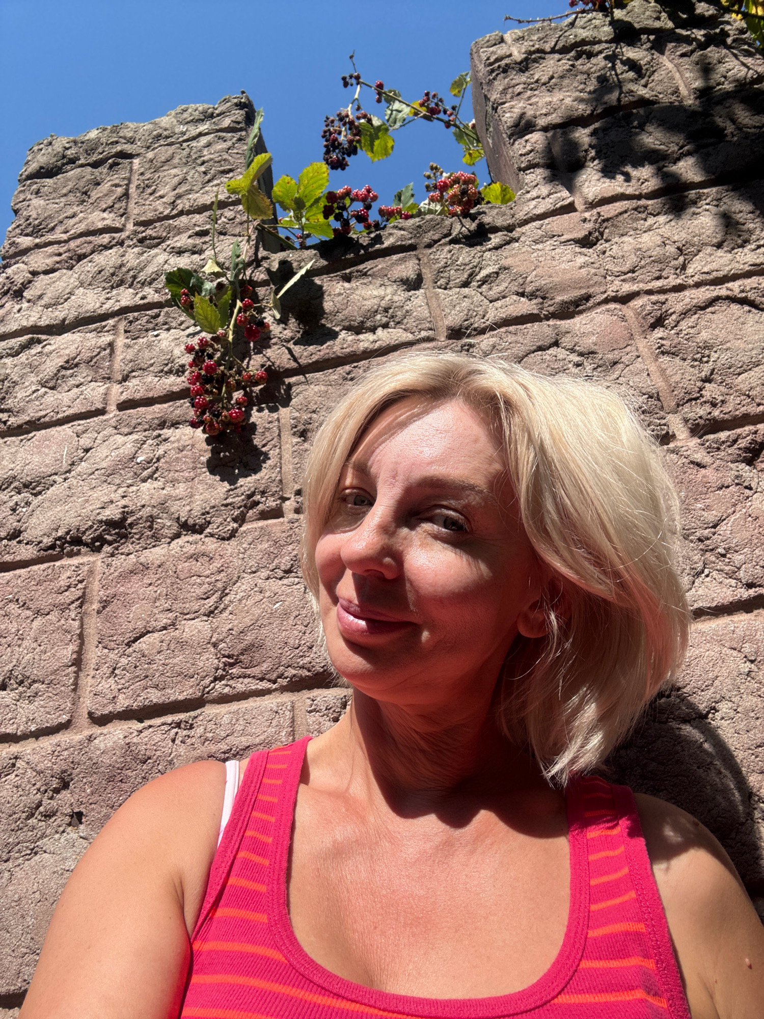 The image shows a woman with blonde hair and fair skin, wearing a pink and orange striped tank top. She is leaning against a rough, textured stone wall. The wall appears to be ancient or historical, with visible mortar lines and a rough-hewn texture. Above and behind her, there is a section of the wall with an opening, through which green leaves and clusters of red and black berries can be seen. The sky is clear and blue. The lighting suggests it is a sunny day, casting shadows on the wall and the woman. The scene is set in Abrau-Dyurso, Russia, a location known for its vineyards and natural beauty, including historical remnants.