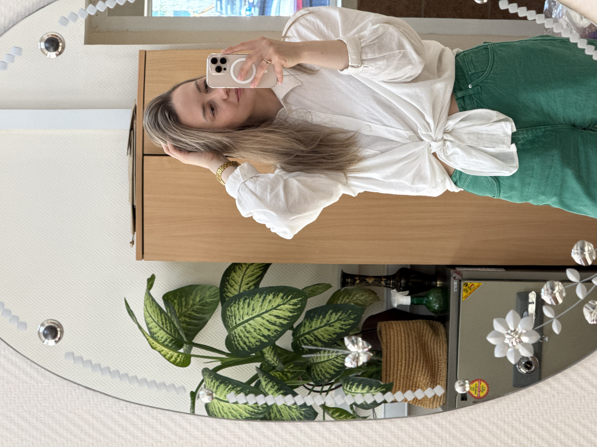 A woman is taking a selfie in a mirror. She is wearing a white blouse tied at the waist and green jeans. The background includes a light brown cabinet, a plant with green and yellow leaves, a safe, and some bottles. The mirror is oval with decorative embellishments. The location is Chaikovsky, Russia.