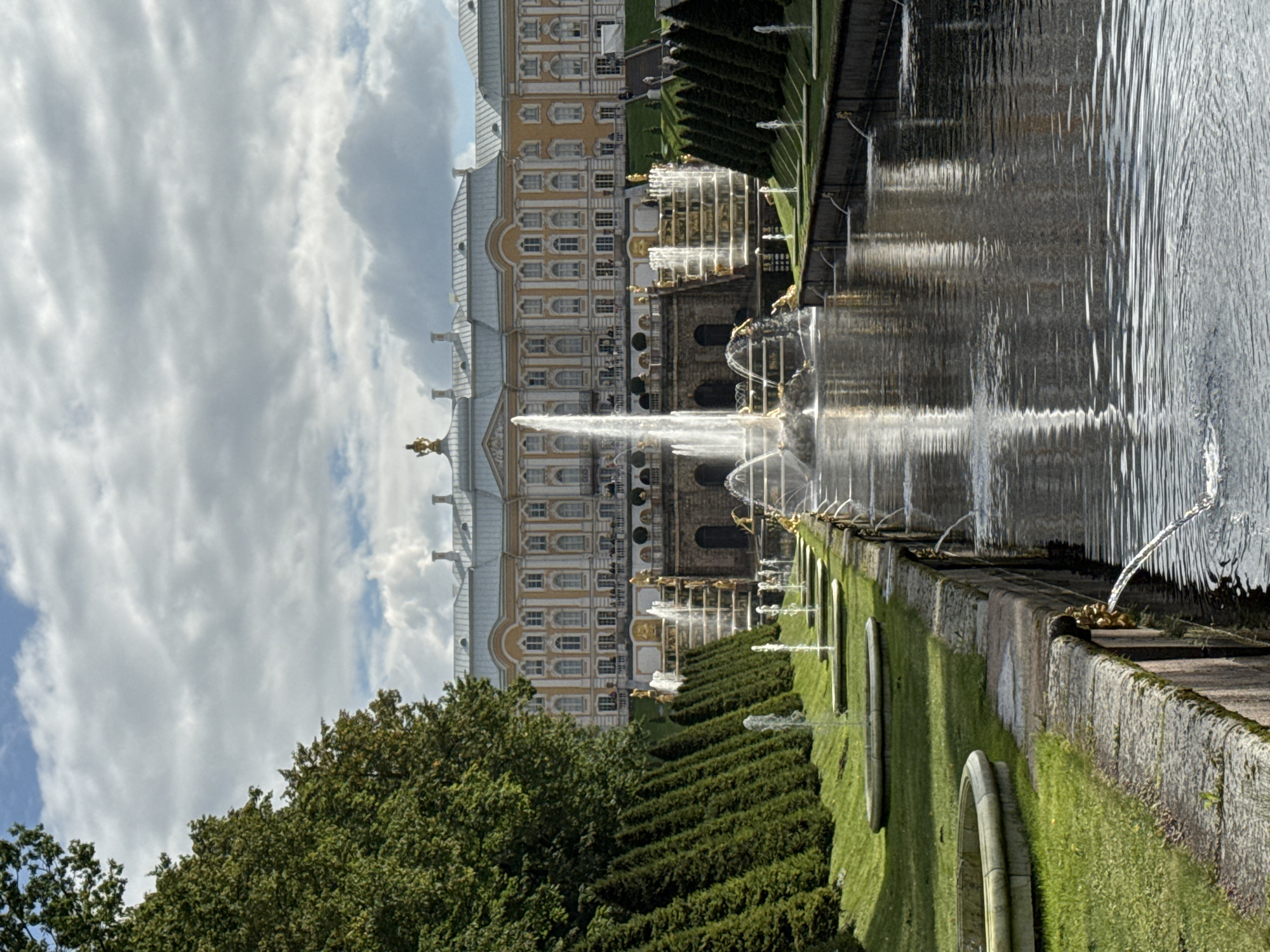 The image shows the Grand Cascade at Peterhof Palace, located in Peterhof, Russia. The Grand Cascade is a spectacular series of waterfalls and fountains, with the central and largest fountain, Samson, at its apex. Samson is depicted as a sculpture of the biblical hero Samson tearing open the jaws of a lion, symbolizing Russia's victory over Sweden in the Great Northern War.
The palace itself, with its distinctive yellow facade and numerous windows, serves as a grand backdrop to the cascade. The architecture is baroque, characterized by its opulence and grandeur. Surrounding the cascade are manicured lawns, meticulously trimmed hedges, and a reflective pool that mirrors the sky and the palace. The sky is partly cloudy, with dramatic clouds casting shadows and highlights on the scene. A few people can be seen on the terraces of the palace and near the fountains, observing the spectacle. The overall impression is one of magnificent natural and architectural beauty, a testament to the imperial splendor of Russia.
