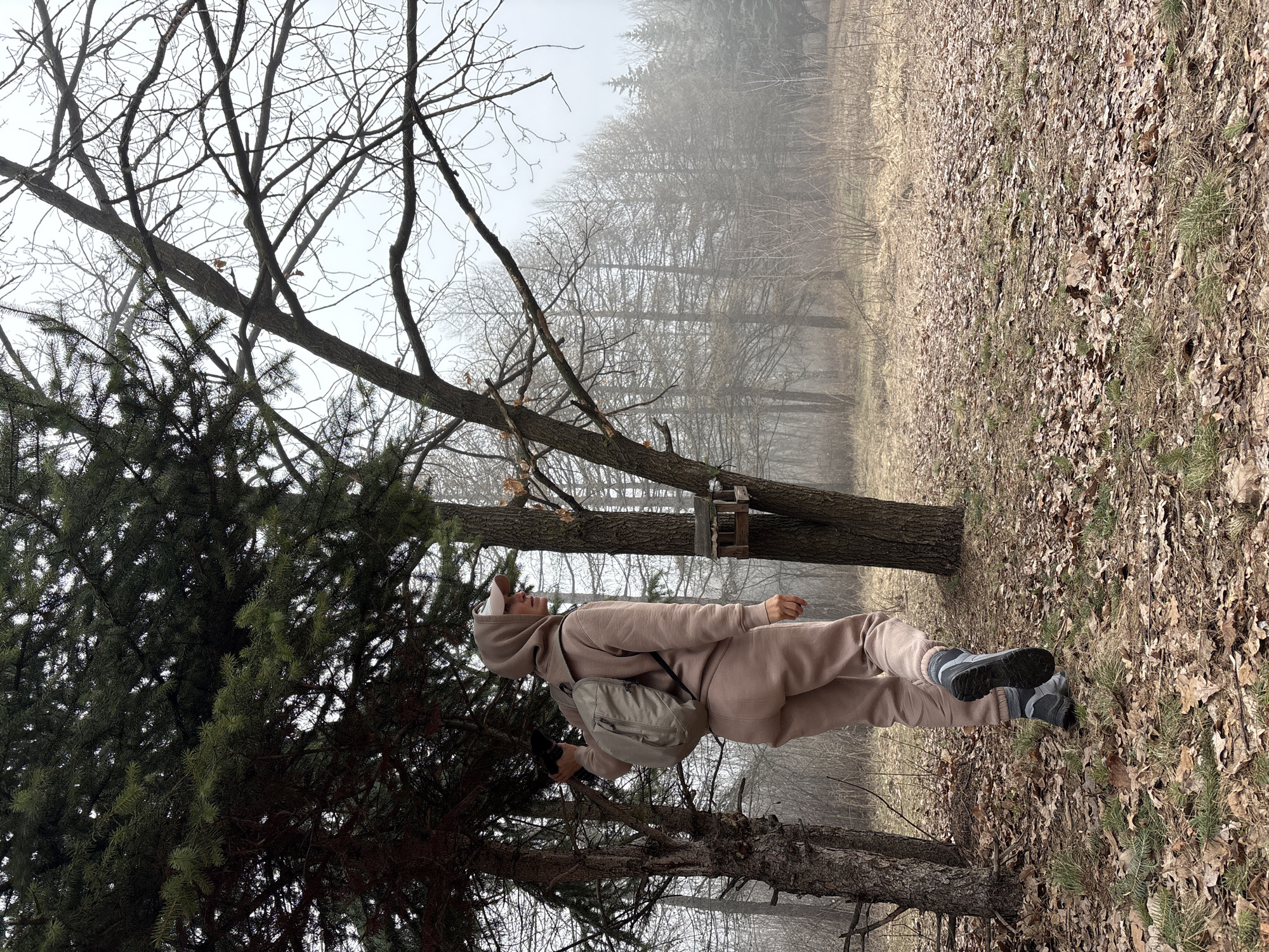 A woman wearing a tan hooded sweatshirt and matching sweatpants stands in a forest setting. She has on a white cap and a backpack. The ground is covered in fallen leaves and there are numerous trees, some bare and some with green foliage. The background is hazy and suggests a foggy or overcast day. The location is likely a park or wooded area within Ulyanovsk, Russia.