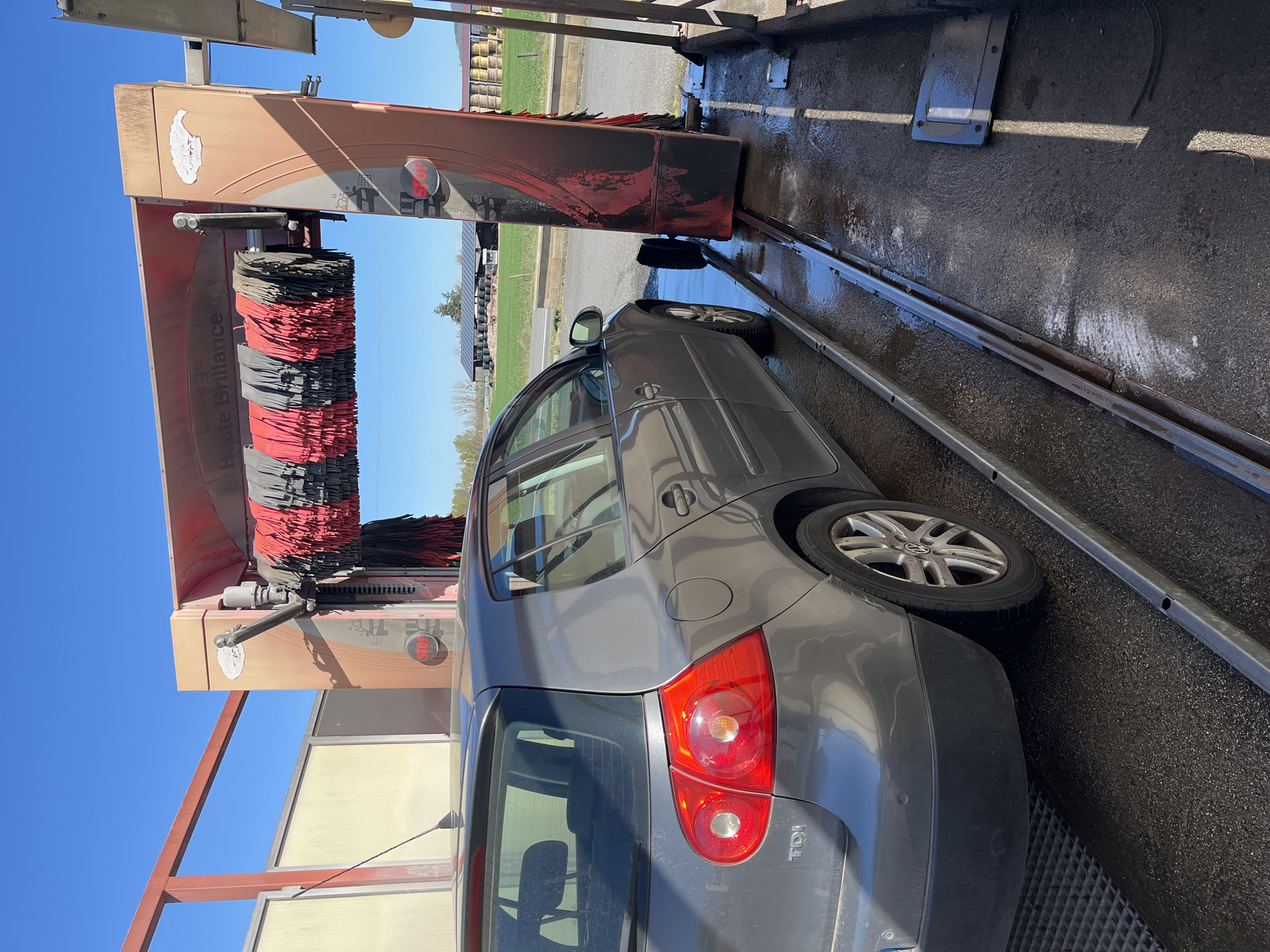 A gray Volkswagen Golf TDI is positioned inside an automated car wash. The car wash features red and black rotating brushes. The sky is clear and blue. The location is Chambœuf, France.