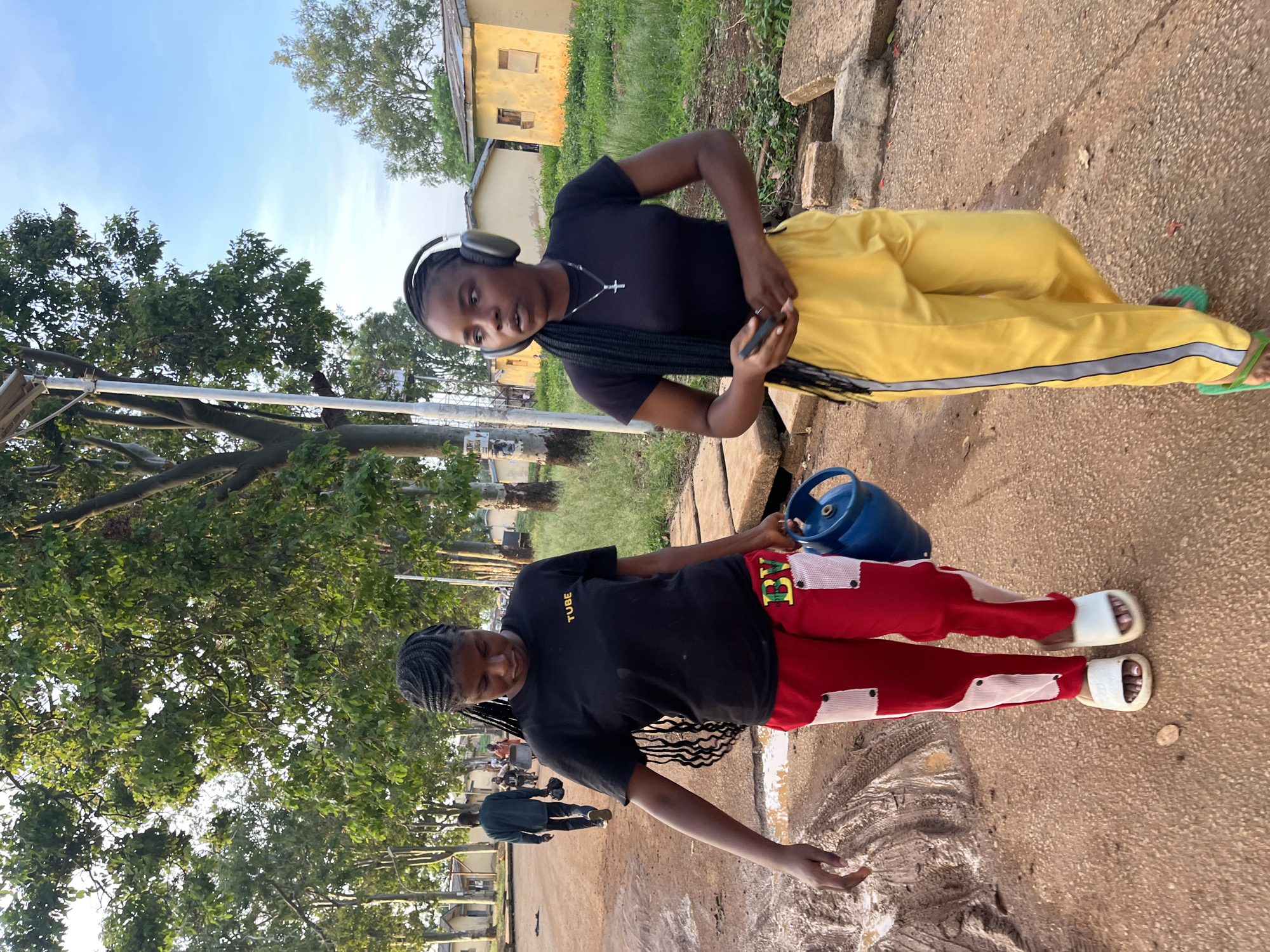 This image, taken in Rafin Par, Nigeria, captures two young women outdoors, likely in the late afternoon or early evening given the soft lighting. The ground is a mix of dirt and concrete, with lush green trees and foliage dominating the background and parts of the foreground. Distant, simple yellowish buildings are also visible.
On the left side of the upright image, a woman with braided hair wears a black t-shirt and red pants with white and black patterned patches. Her t-shirt has the word "TUBE" printed in yellow text, and her red pants feature "BV" in yellow with a green outline on one of the patches. She is wearing white slides/slippers and is carrying a blue gas cylinder by its handle, suggesting she is transporting it. Her gaze is slightly downwards, focused on her activity.
On the right, another woman with braided hair wears a black t-shirt, yellow pants with a grey side stripe, and green flip-flops. She is wearing white headphones and has a cross pendant on her necklace. She holds a mobile phone in one hand and looks towards the camera with a neutral expression.
In the background, another person in dark clothing is partially visible. The scene depicts a casual, everyday outdoor activity in a semi-urban or rural setting.