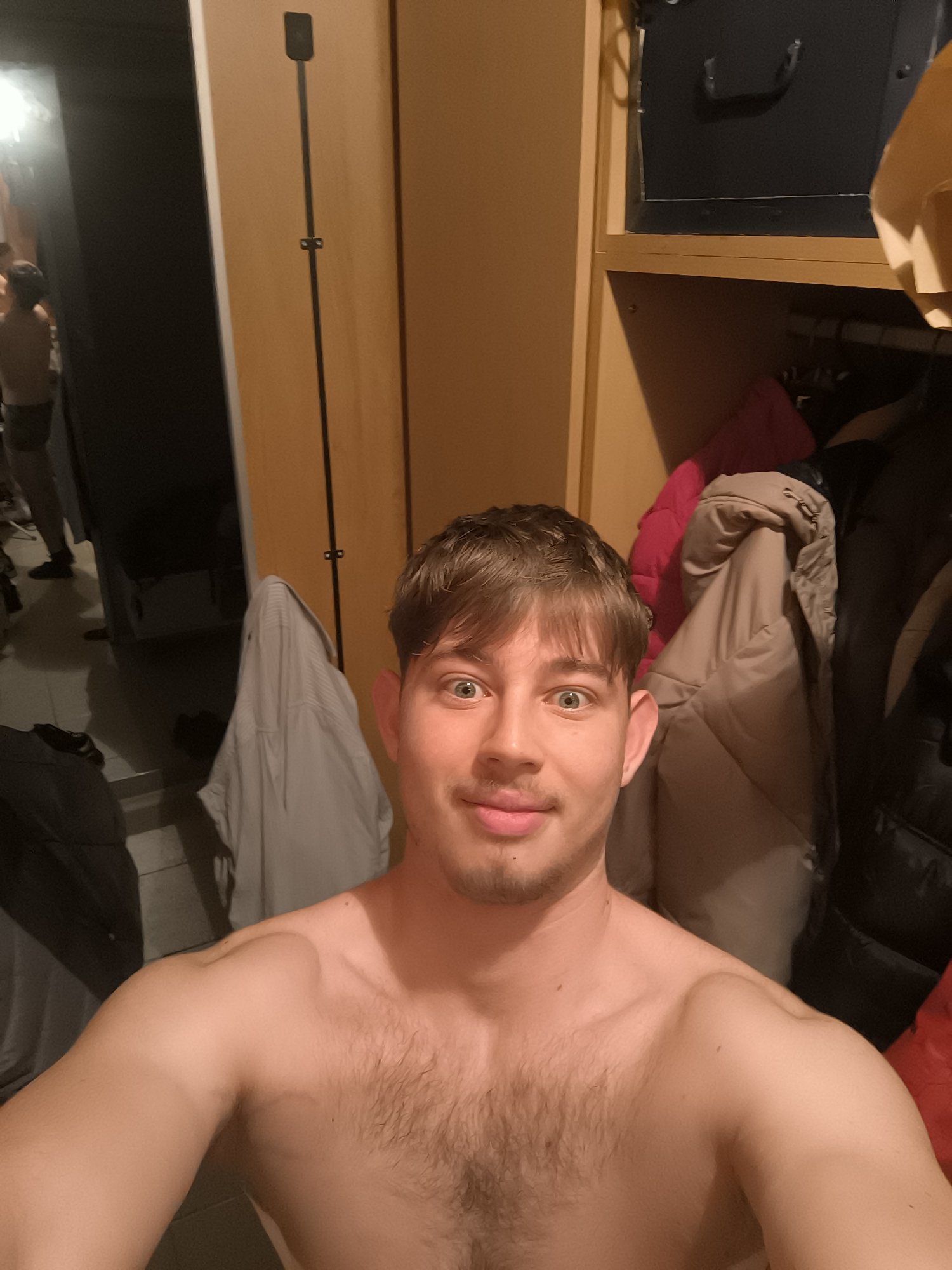 A selfie of a shirtless man with light brown hair and green eyes, positioned in what appears to be a hallway or closet space. The man is smiling with a slight stubble beard. The background shows a light-colored door or wall panel, a hanging rack with various coats and garments, and potentially a shelf unit with items on top. A second individual partially visible in a mirror can be seen in the far background. The location is unknown and does not provide details or context allowing for the conclusion to be in Sofia, Bulgaria.