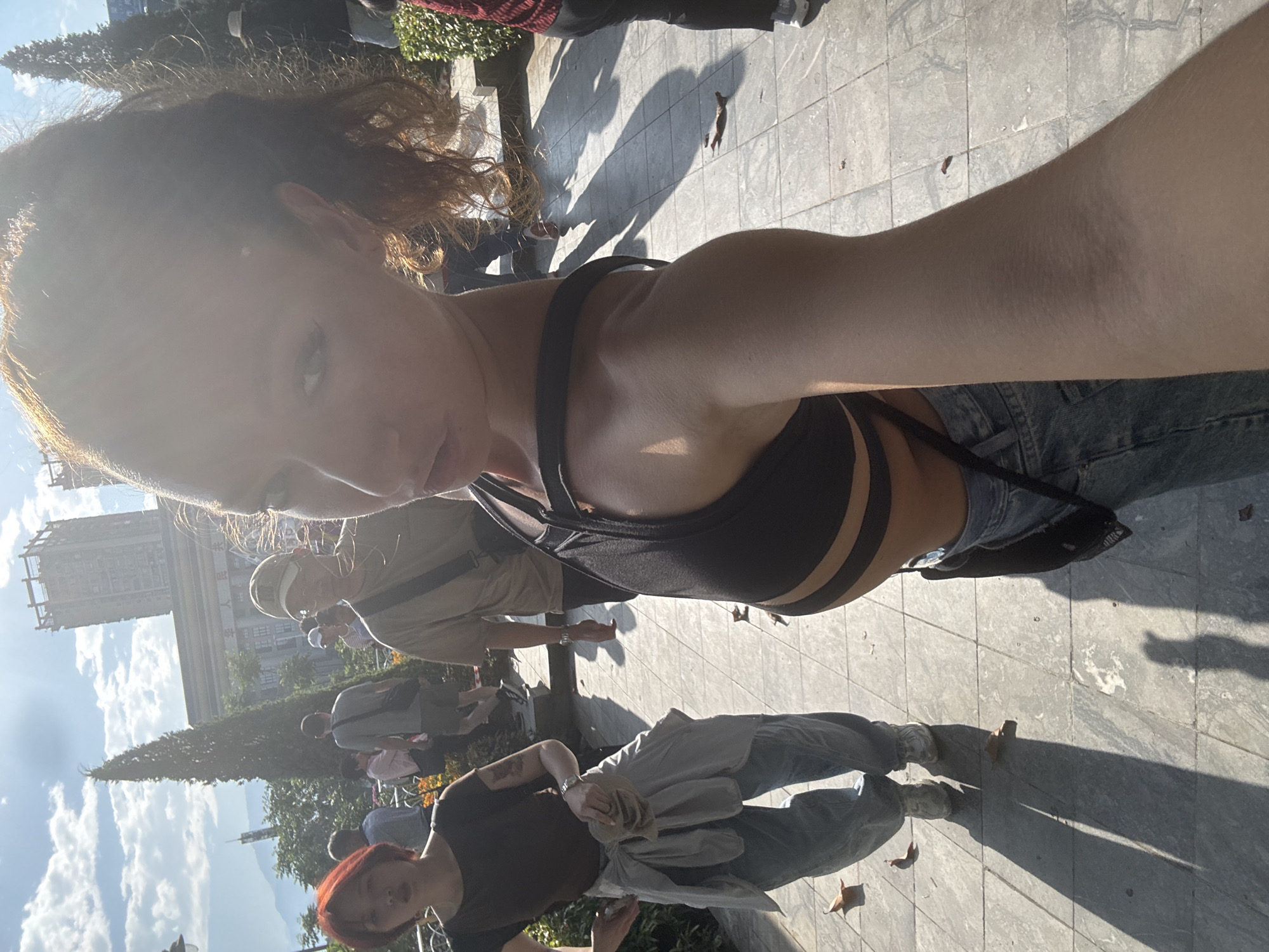A selfie taken outdoors in Lao Cai Ward, Vietnam. A woman with light skin, curly brown hair, and a dark sports bra is visible in the foreground. The background includes people walking on a paved surface with trees and buildings visible in the distance. The weather appears sunny, casting shadows.