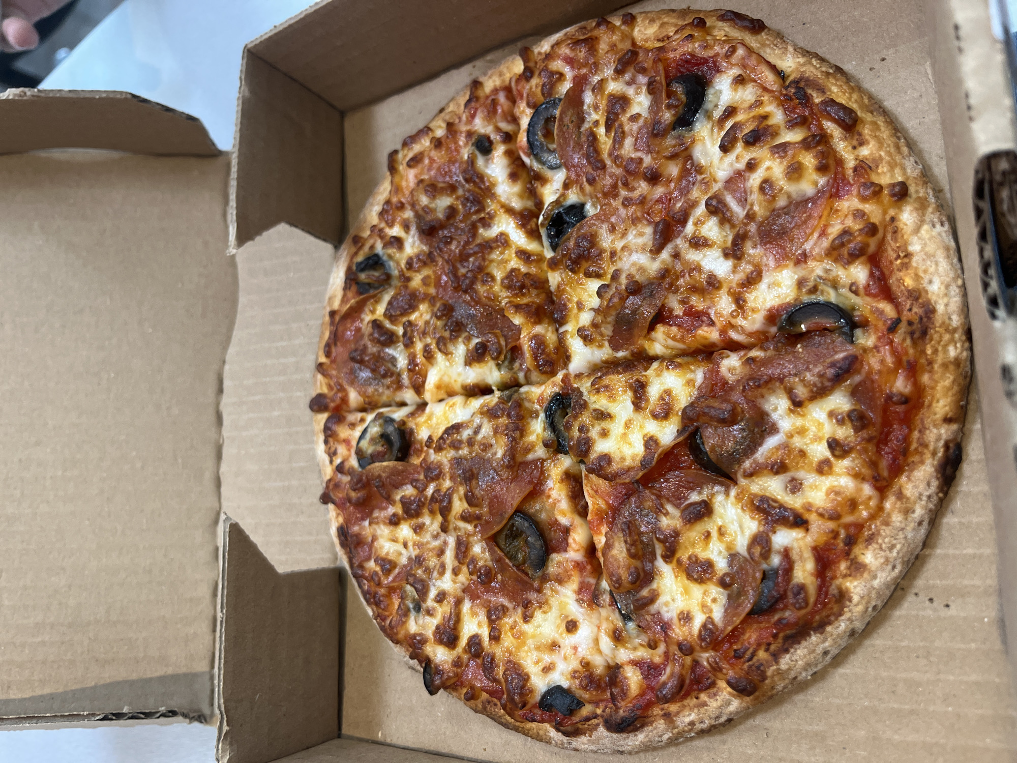 The scene depicts a whole, circular pizza, pre-cut into eight slices, resting inside an open, light brown corrugated cardboard pizza box. The pizza features a golden-brown crust, a tomato sauce base, and is topped with an even layer of melted, bubbly, and slightly browned mozzarella cheese. Distributed across the cheese are numerous reddish-orange, curled pepperoni slices and dark, circular black olive slices. The interior of the cardboard box, with its textured surface, forms the immediate environment for the pizza. Beyond the box, a light-colored surface, possibly a tabletop, is partially visible, along with a blurry, lighter background. The fingers of an individual are faintly discernible in the upper right section of the background. This scene is located in San Bernardino Tlaxcalancingo, Mexico.