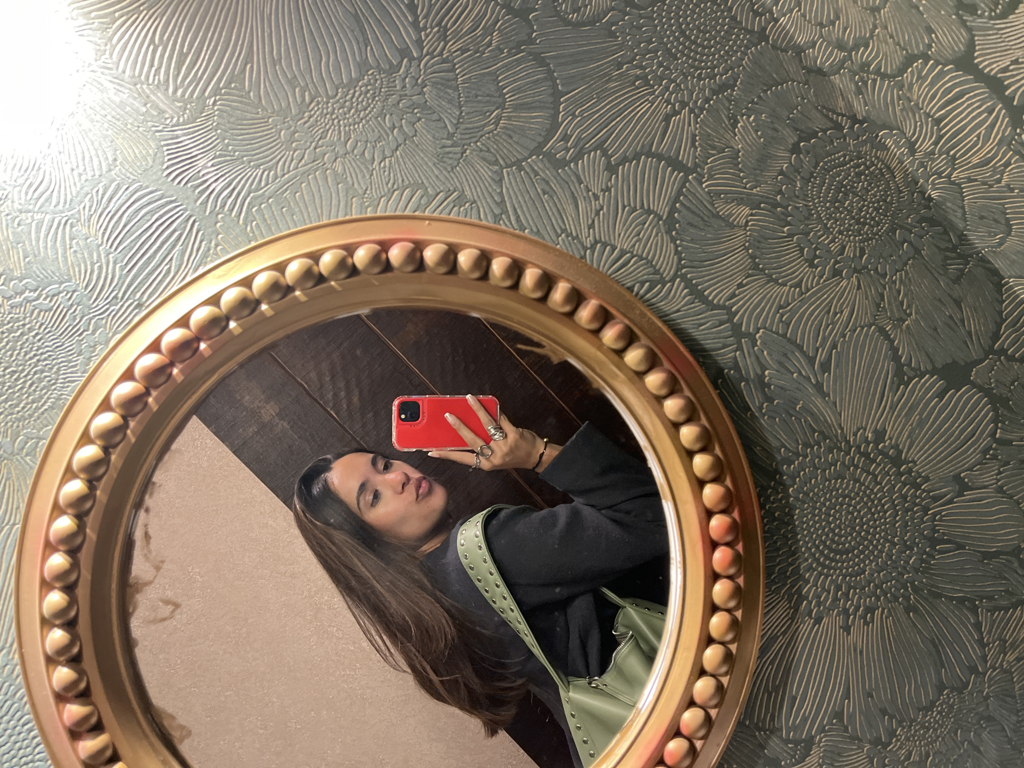 A woman is taking a selfie in a round gold-colored mirror. The mirror hangs on a wall covered with patterned wallpaper in shades of blue and gray, resembling a stylized floral design. The mirror's reflection shows the woman holding a red smartphone. She has long, dark hair and wears a black jacket. A green purse hangs from her shoulder. The background in the mirror's reflection reveals a wooden wall and a beige wall. The location cannot be ascertained specifically to San Bernardino Tlaxcalancingo, Mexico, from the visual information.