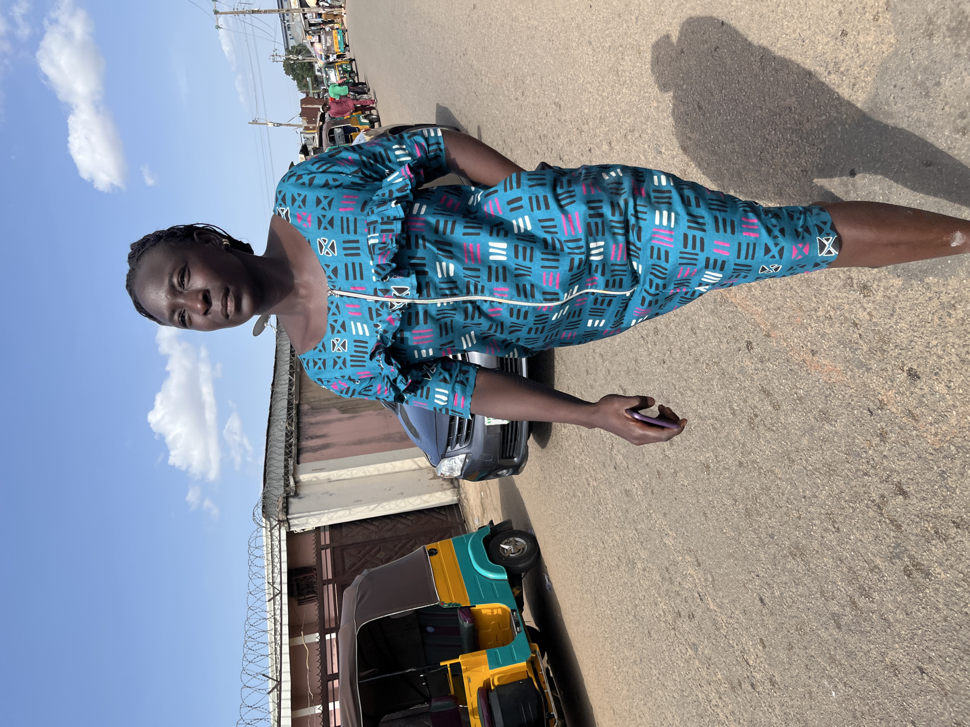 A woman with braided hair walks down a street in Jos, Nigeria. She is wearing a teal and white patterned dress with a zipper down the front and ruffled trim. She holds a purple smartphone in her right hand. In the background, a yellow and teal auto-rickshaw is parked, and a gray car is driving down the street. The sky is blue with scattered clouds. The street appears to be lined with buildings and overhead wires. The woman's shadow is cast on the asphalt road.