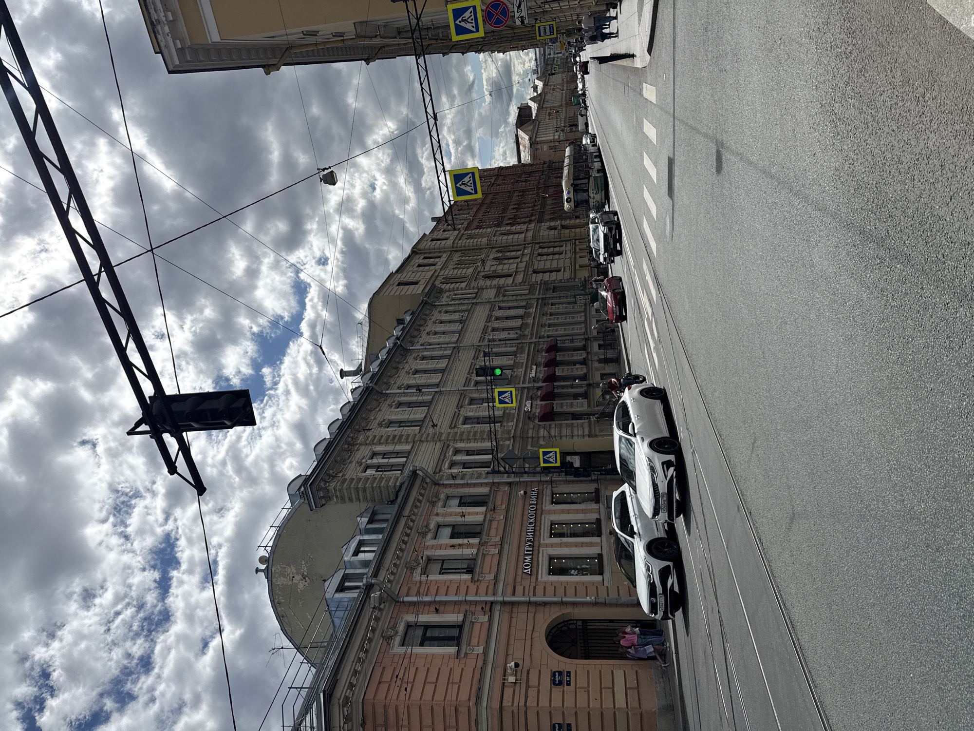 The image shows a view of a street in Saint Petersburg, Russia, on a cloudy day. The street is wide with tram tracks. Several cars are visible, including a white sedan and a white SUV parked near a building. A bus is also present further down the street. Pedestrians are walking on the sidewalk. Buildings line both sides of the street, with architectural details typical of Saint Petersburg. There are traffic lights and pedestrian crossing signs visible. The sky is filled with dramatic clouds. The scene appears to be an urban street with active traffic.