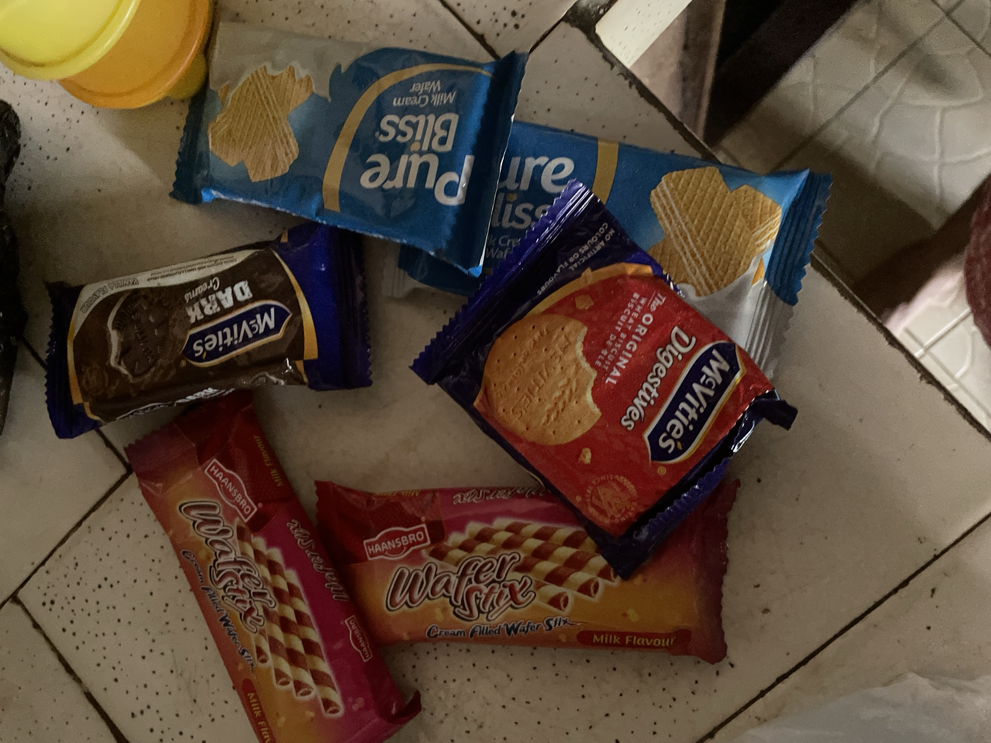 The image shows a pile of assorted biscuits and wafers on a tiled floor. There are at least five visible packets of biscuits. Two packets are for "Wafer Stick" brand, one with a red and yellow wrapper, and another with a predominantly red wrapper. Both advertise "Milk Flavour" and "Cream Filled Wafer Stick". A dark brown packet of "McVitie's Dark Chocolate Cream" biscuits is also visible. Two blue packets of "Pure Bliss Milk Cream Wafer" are present, with one partially obscured. A red packet of "McVitie's Digestive" biscuits is also visible, showing text in English. The environment appears to be indoors with a tiled floor, suggesting a domestic or commercial setting. The lighting is dim, possibly from overhead artificial light. No people are visible in the image. The location is described as Kakau, Nigeria.