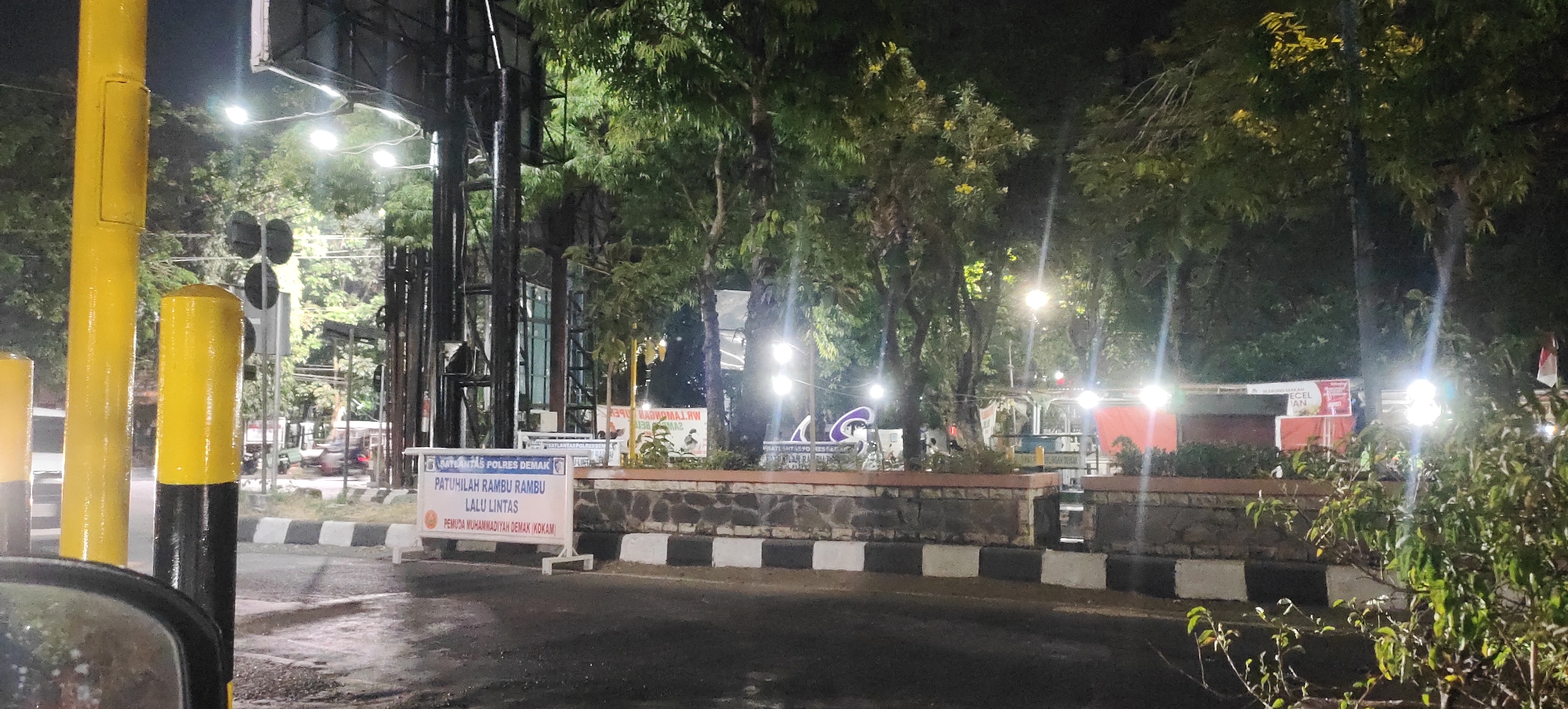 The image shows a street scene at night in Demak, Indonesia. In the foreground, a yellow and black striped bollard is visible on the left. Behind it, a white sign with blue and red text, bearing the emblem of "SATLANTAS POLRES DEMAK" and the words "PATUHILAH RAMBU RAMBU LALU LINTAS" and "PEMUDA MUHAMMADIYAH (KOKAM)", is prominently displayed. A low stone wall with black and white checkered patterns separates the sidewalk from the road. Several trees with illuminated foliage are present in the background, with streetlights casting a glow. Behind the trees, there are structures that appear to be stalls or buildings with red and white banners. In the distance, vehicles are parked or in motion on the road, and there are various poles and overhead structures, suggesting an urban environment. The overall impression is of a street corner or an intersection at night, possibly near a police station or a public gathering area.