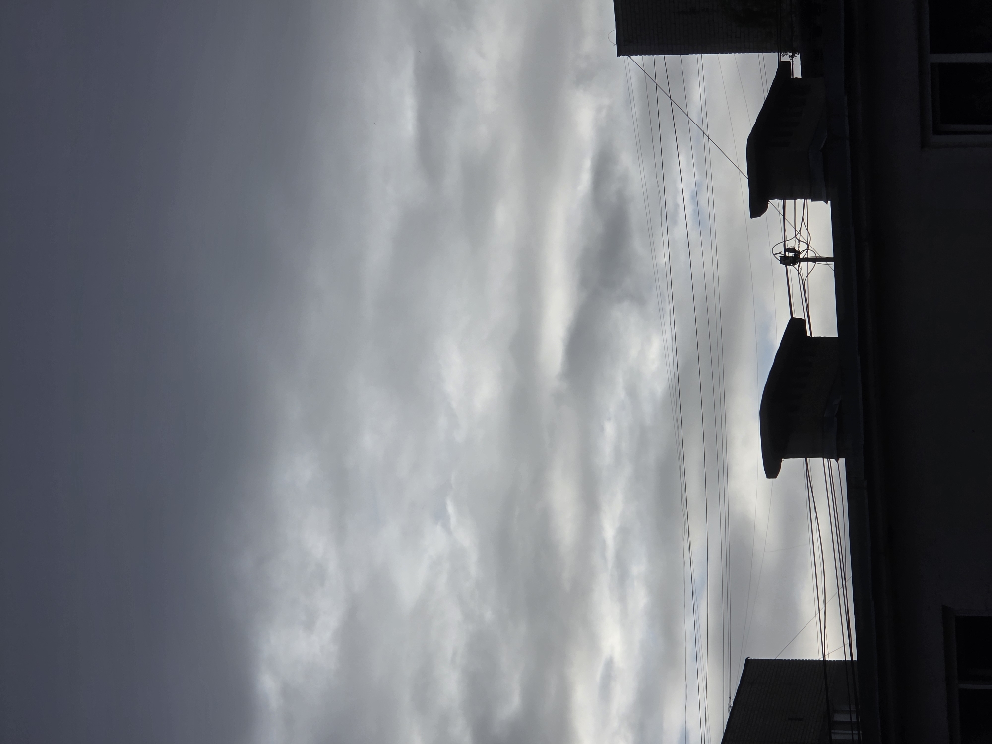 The image depicts a cloudy sky above the rooftops of buildings in Chaikovsky, Russia. The sky is filled with various shades of gray clouds, with some areas appearing brighter where light filters through. Silhouetted against the sky are the dark shapes of building rooftops, including two distinctive ventilation caps or chimneys on the central rooftop. Power lines crisscross the frame, further silhouetted against the cloudy backdrop. On the far left and far right of the image, parts of brick buildings are visible. The overall scene is monochromatic and somber, suggesting overcast weather. There are no people or animals visible in the image.