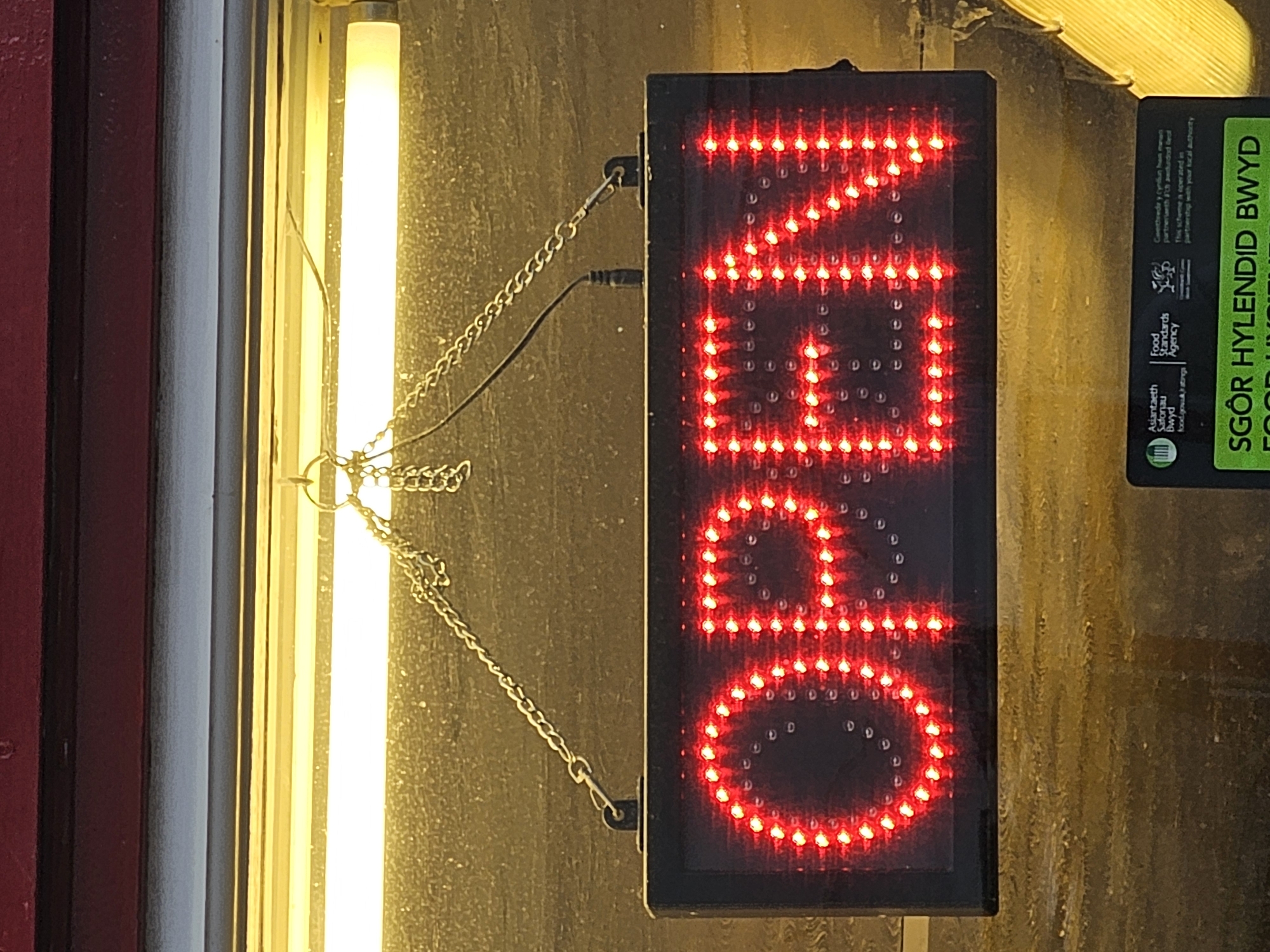 The image shows an "OPEN" sign hanging from chains in Holywell, United Kingdom. The sign is a black rectangular box with the word "OPEN" displayed vertically in bright red LED lights. To the left of the sign, a fluorescent light emits a bright yellow glow. To the right, a food hygiene rating sticker is partially visible. The background appears to be a textured surface, possibly a wall.