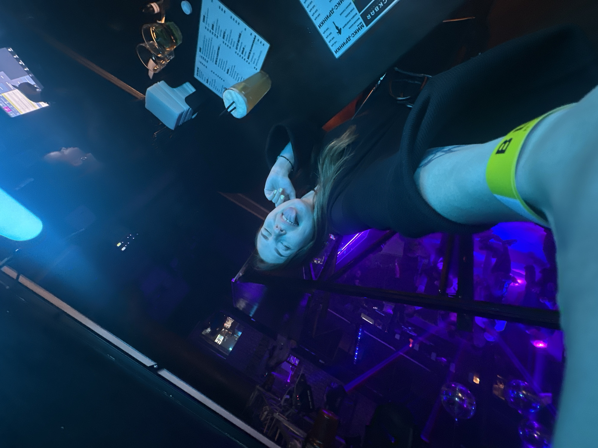The image shows a woman in a dimly lit nightclub, likely in Perm, Russia, based on the Cyrillic text on a menu visible in the foreground. The woman is leaning back, with her tongue sticking out, and appears to be taking a selfie. She is wearing a dark outfit and a bright yellow wristband. In the background, there are disco balls, purple and blue lights, and other patrons. On the table in front of her are two glasses, a small stack of white napkins, and a menu. Another menu with Cyrillic text is visible on the right side of the frame. The overall atmosphere is lively and energetic, typical of a nightclub setting.
