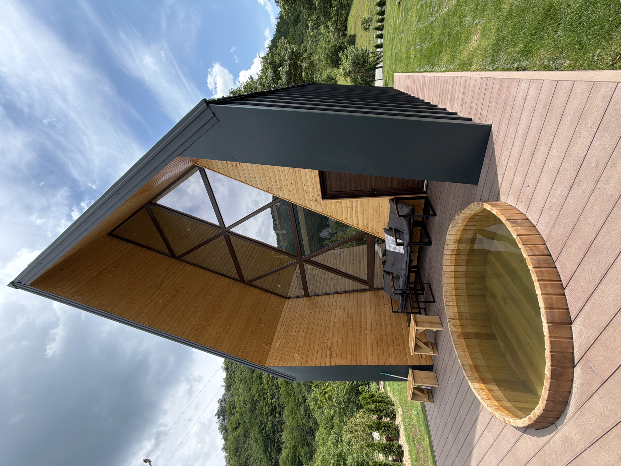 In Bistrica, Montenegro, a modern, angular wooden and dark-paneled house sits on a deck overlooking a grassy slope and a lush, tree-covered mountainside. In the foreground, a large, oval wooden hot tub filled with murky water is situated on the deck. The house features a striking triangular facade with a large, geometric window and a brown wooden door. On the deck, a dark-colored sofa with grey cushions and a white checkered throw is placed near two small, light-wood stools. The environment is characterized by bright daylight under a partly cloudy sky.