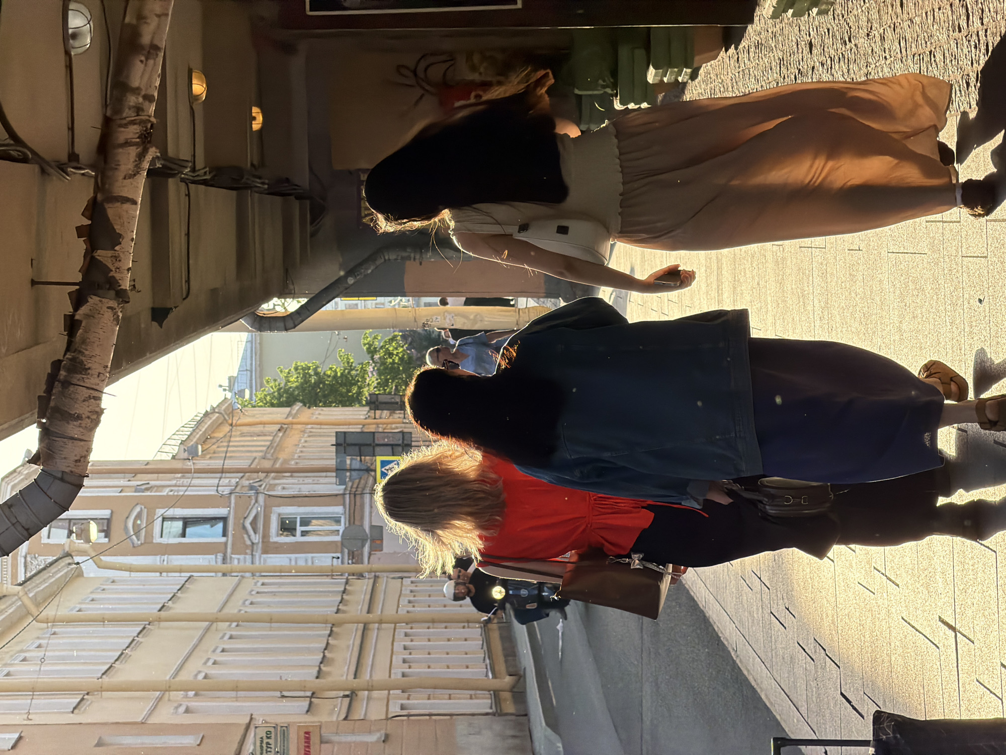 The image depicts a street scene in Saint Petersburg, Russia. Three women are walking away from the camera on a brick sidewalk that is lit by direct sunlight. The first woman is wearing a light colored top, a light brown skirt, and a white fanny pack. The second woman is wearing a red top and black pants with a dark shoulder bag, and a blue denim jacket. The third woman has dark hair, is wearing a blue top, and a long blue skirt. In the background, there are buildings with ornate facades, typical of Saint Petersburg's architecture. A birch tree trunk with peeling bark is visible on the left side of the frame, along with piping on the side of the building. A man wearing a white helmet and riding a scooter is visible behind the third woman.