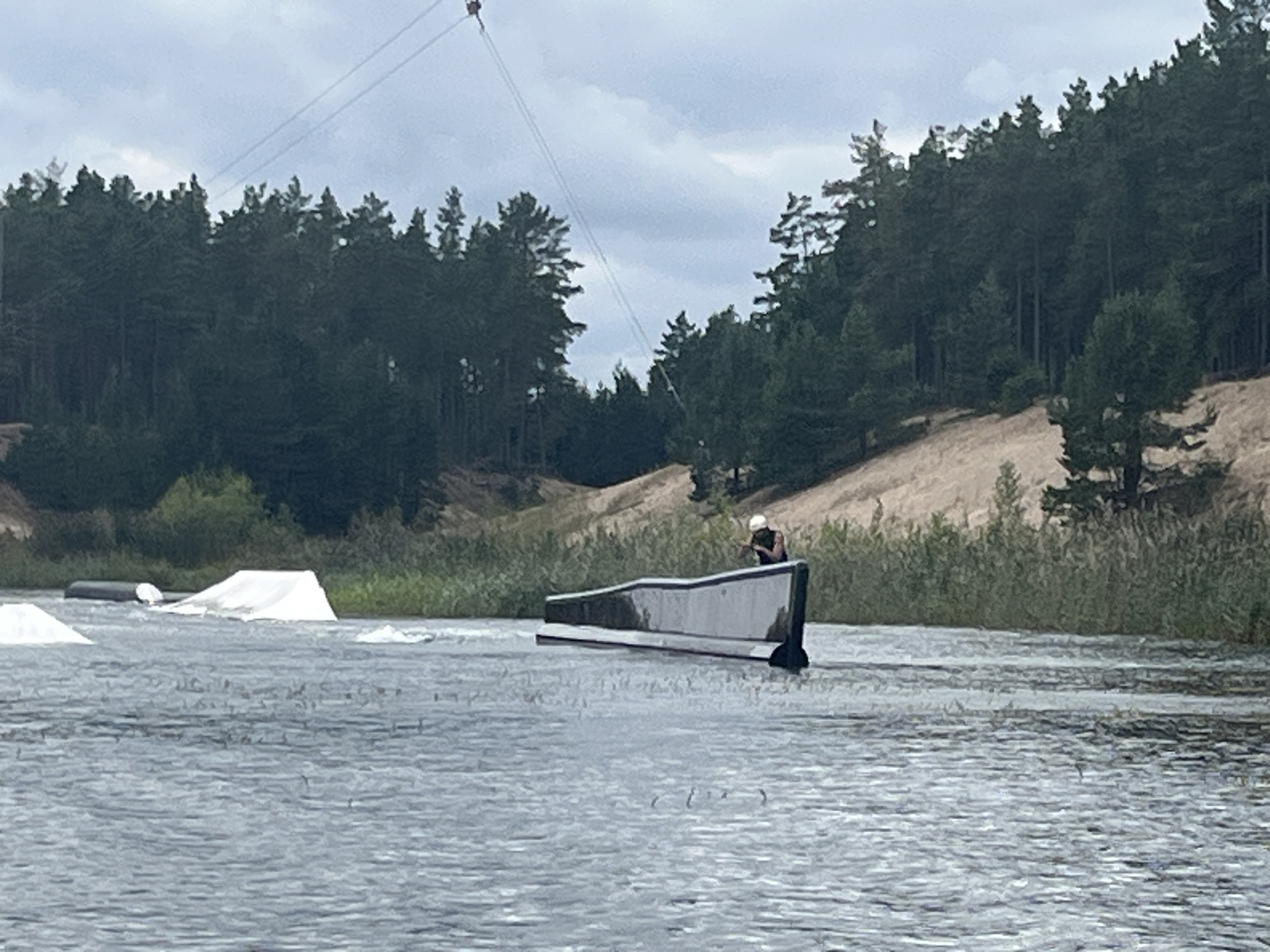 In Männiku, Estonia, a wakeboarding course is depicted on a body of water. A wakeboarder, wearing a white helmet and dark attire, is positioned on a dark, angled ramp that emerges from the water. The ramp is approximately two meters long and one meter high. In the background, a dense forest lines the shore, with a sandy embankment visible behind the trees. The sky is overcast with clouds. Two white, triangular ramps are also present in the water to the left of the main ramp. A cable system, likely for the wakeboarding tow, extends from the upper left corner of the frame.