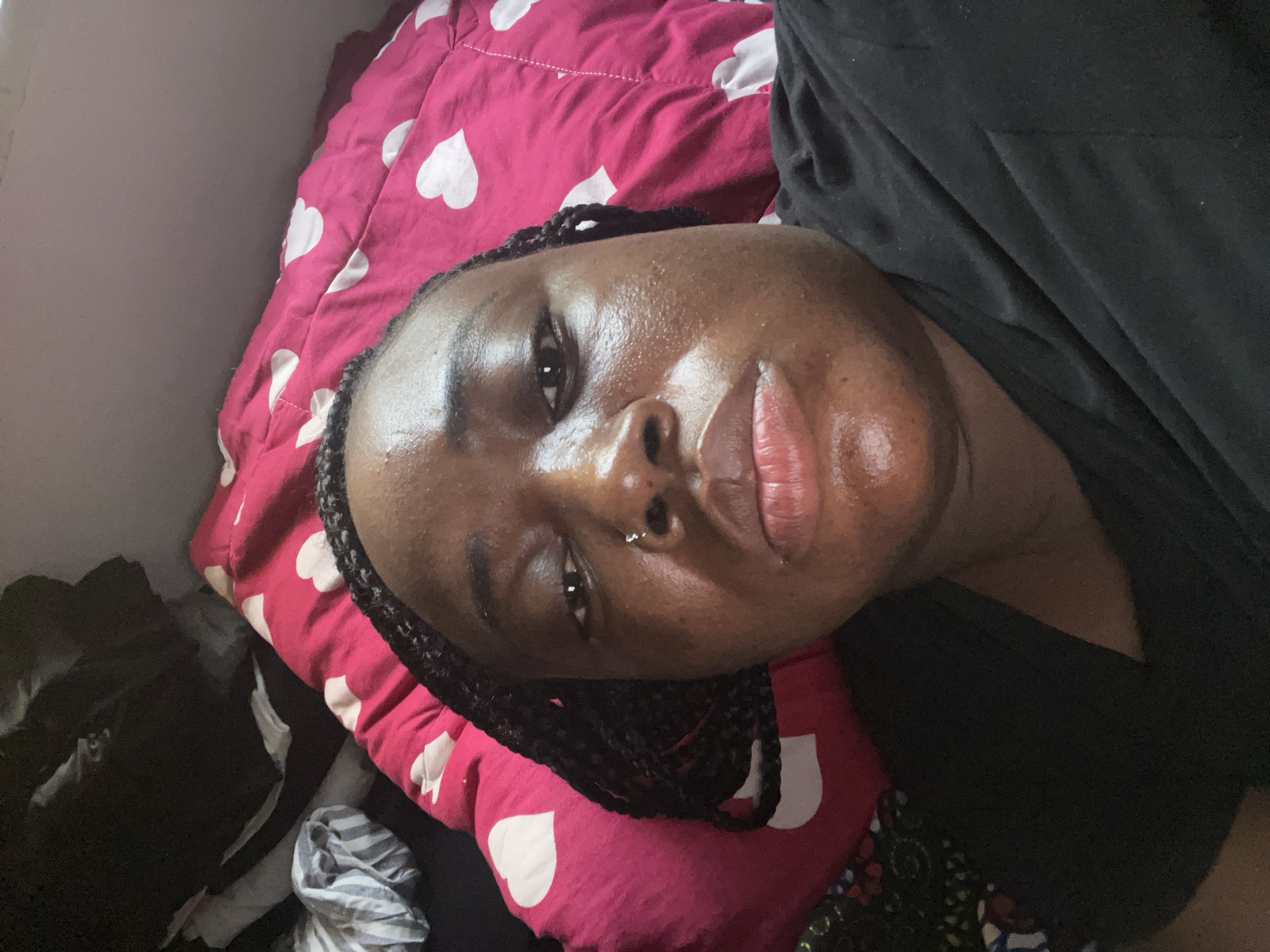 The image is a medium close-up portrait of an individual with dark skin, lying down and looking directly at the camera. The person's head is tilted to the right, suggesting the photo was taken from an overhead or slightly angled perspective, possibly a selfie. They have braided hair framing their face and a small silver hoop piercing in their right nostril. Their skin has a visible sheen, particularly on the forehead, nose, and chin. The individual's lips are full, and their expression appears neutral or relaxed.
They are wearing a black top or t-shirt. The person is resting on a vibrant pink pillow patterned with numerous white hearts. A matching or similar pink and white heart-patterned duvet or blanket is visible behind the pillow. In the lower left corner of the frame, several dark and light-striped fabric items are visible, possibly clothes or other textiles, suggesting a somewhat casual or lived-in indoor environment. The lighting is soft but illuminates the face well, creating highlights on the skin. The setting appears to be an indoor space, likely a bedroom, situated in Abuja, Nigeria. There is no visible text in the image.